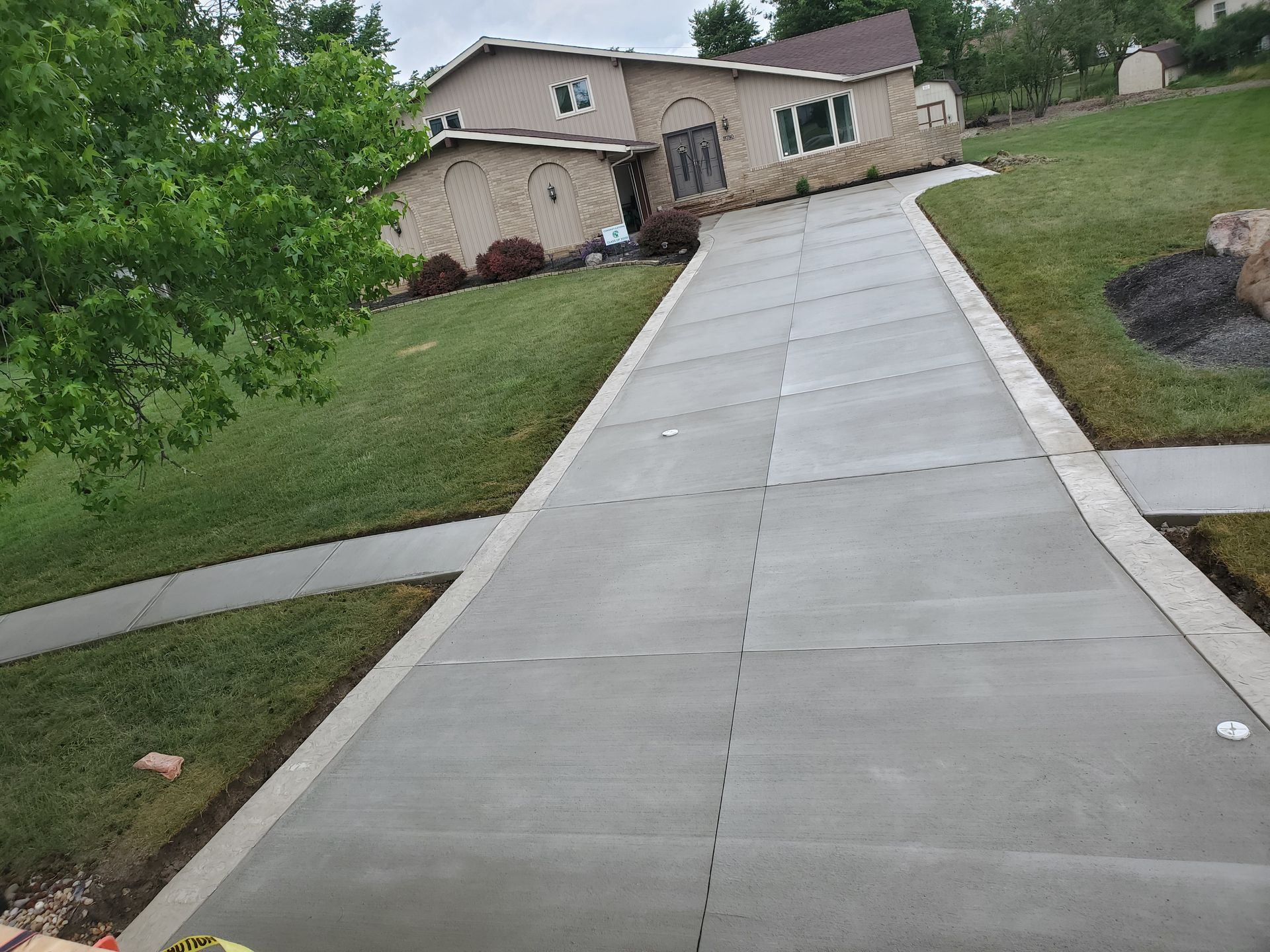 A newly paved, light gray concrete driveway leads up to a tan house with a green lawn on a bright day.