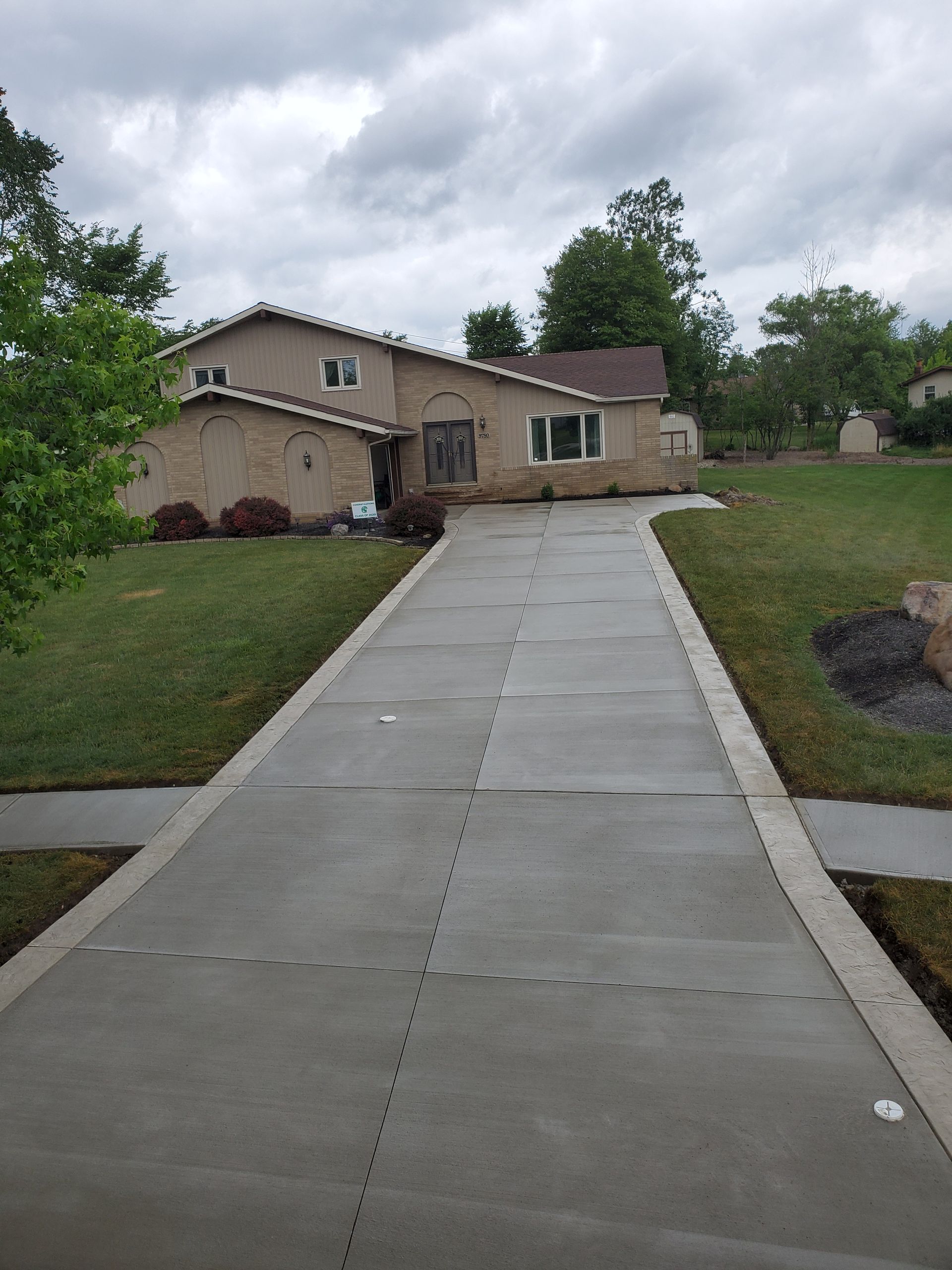 A tan house with a brown roof sits at the end of a long, newly poured concrete driveway lined with a grassy lawn.