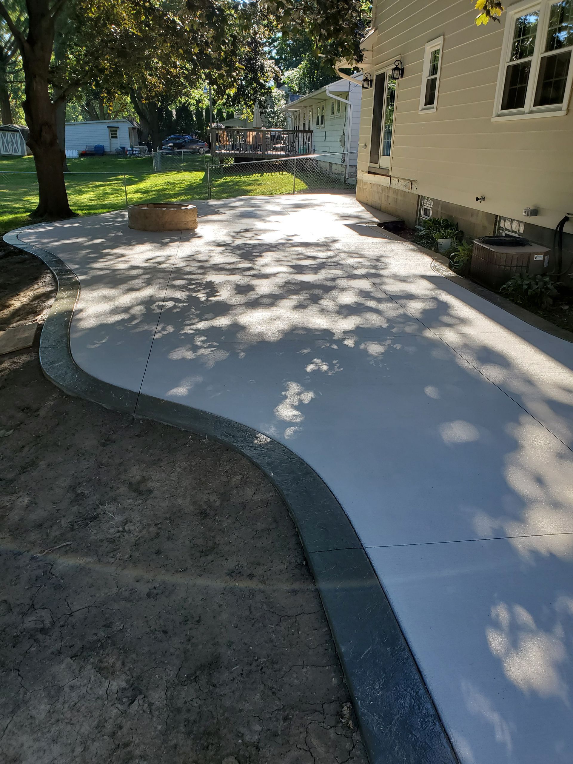 A light-colored, curved concrete patio with a dark speckled border edge, set in a grassy backyard next to a house.