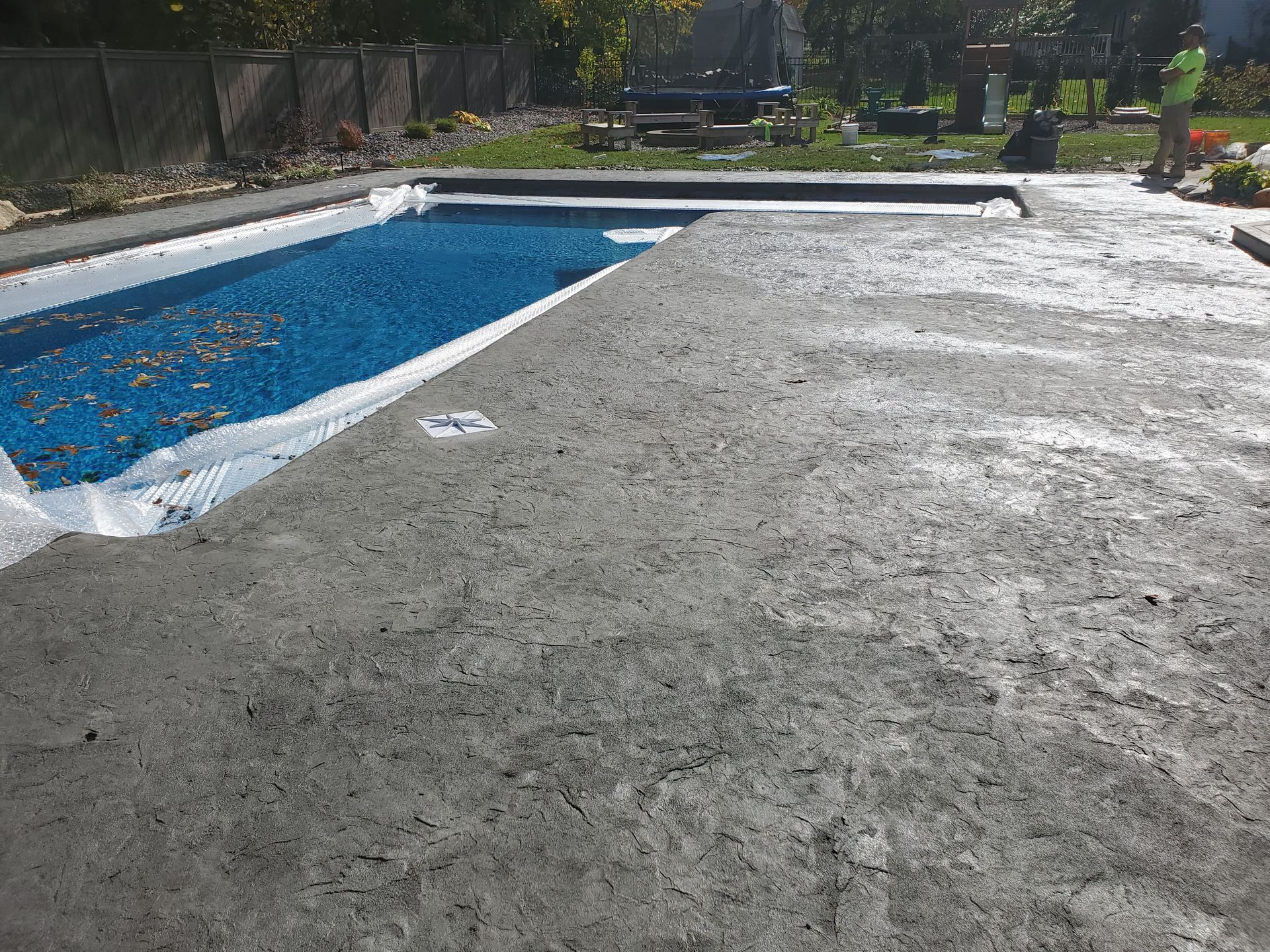 Freshly poured concrete patio surrounding a swimming pool under a blue tarp in a backyard.