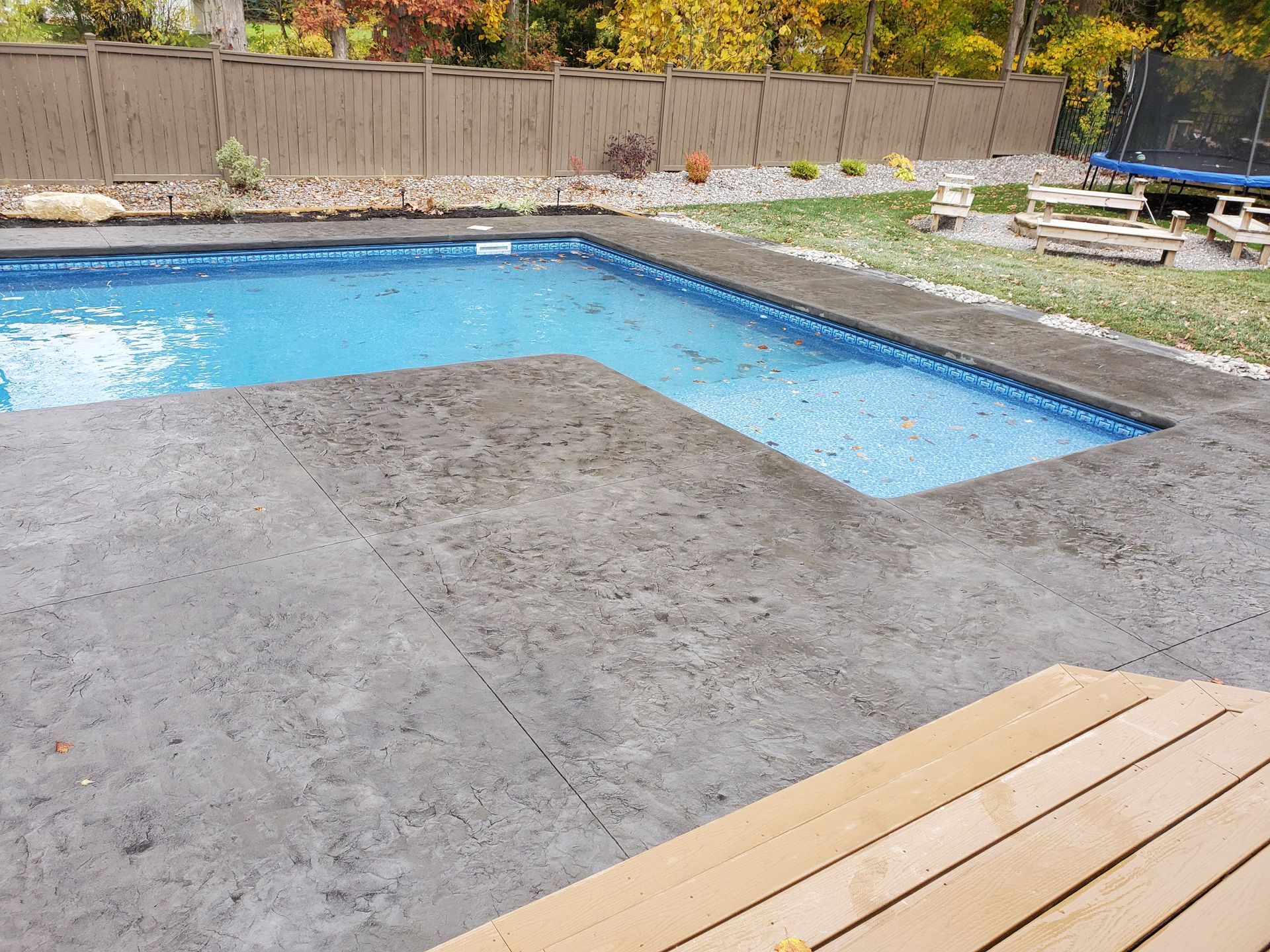 A rectangular swimming pool with blue water sits in a grey stamped concrete patio near a wooden fence and lawn.