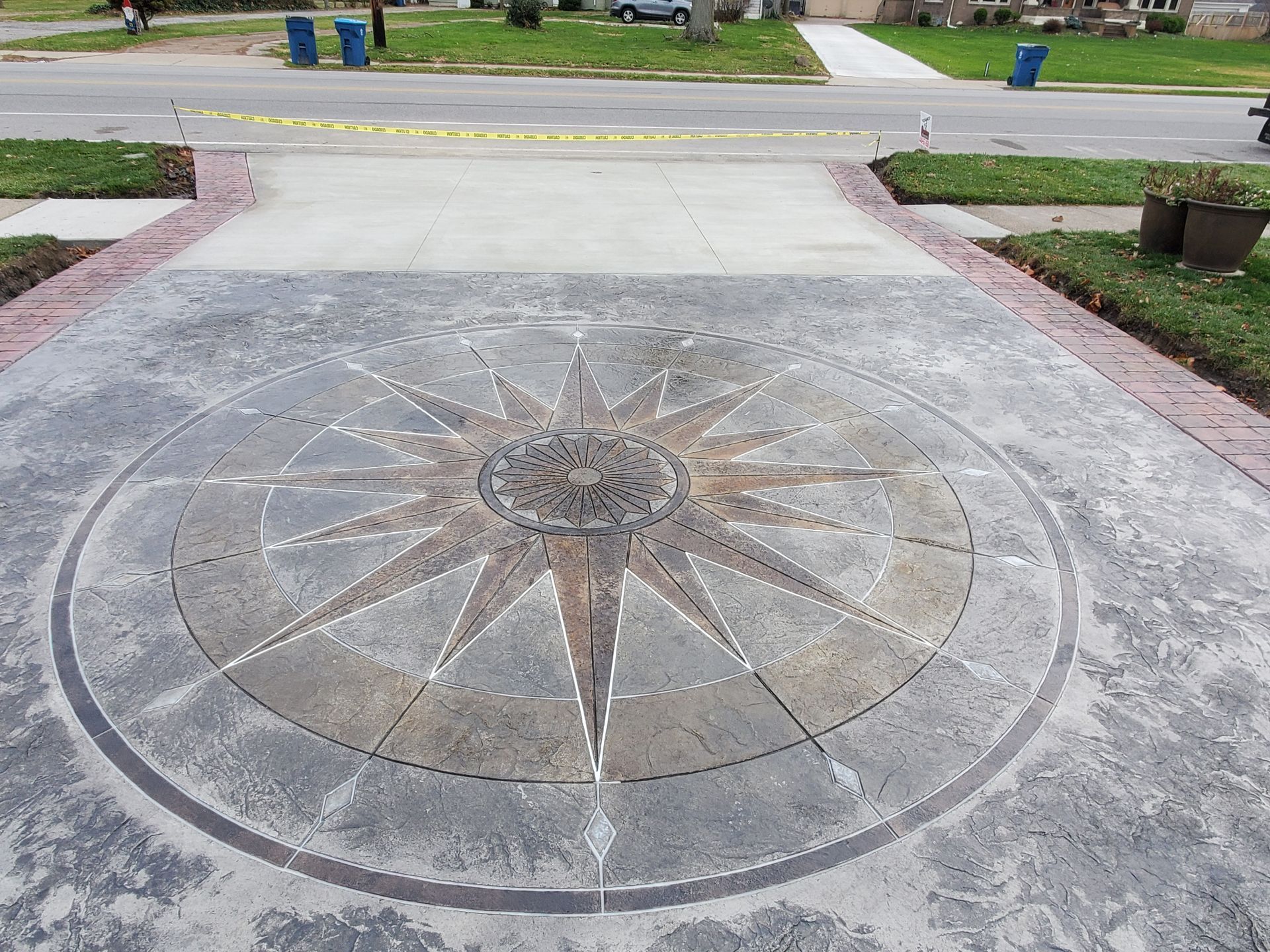 A driveway with a decorative, circular compass-rose inlay in neutral stone tones, bordered by red brick pavers.