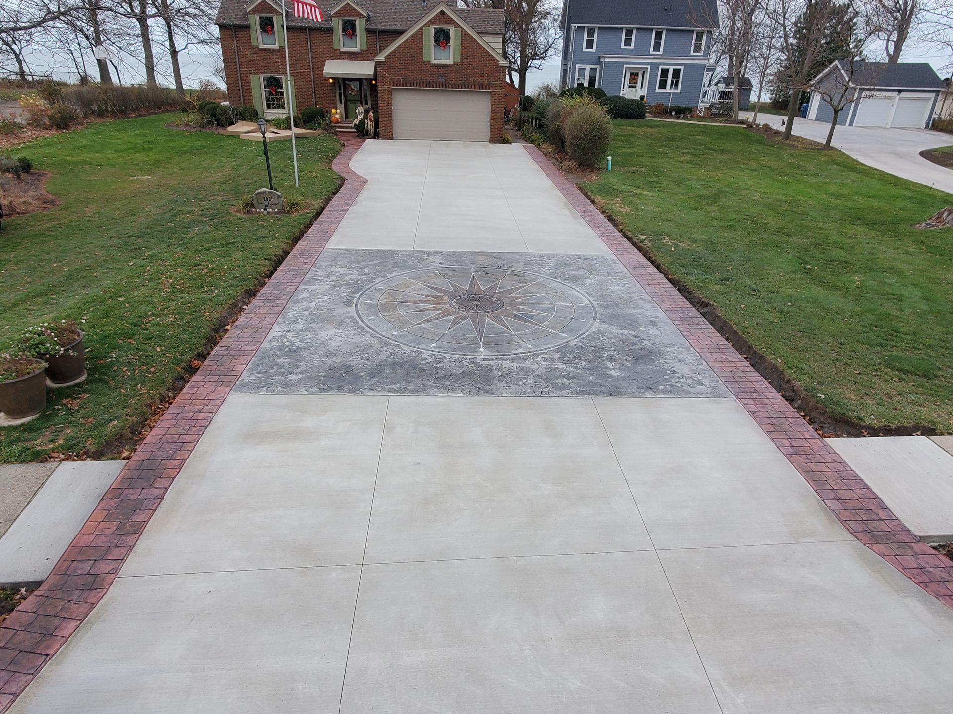 A residential concrete driveway featuring a decorative center medallion and dark red brick border edging.