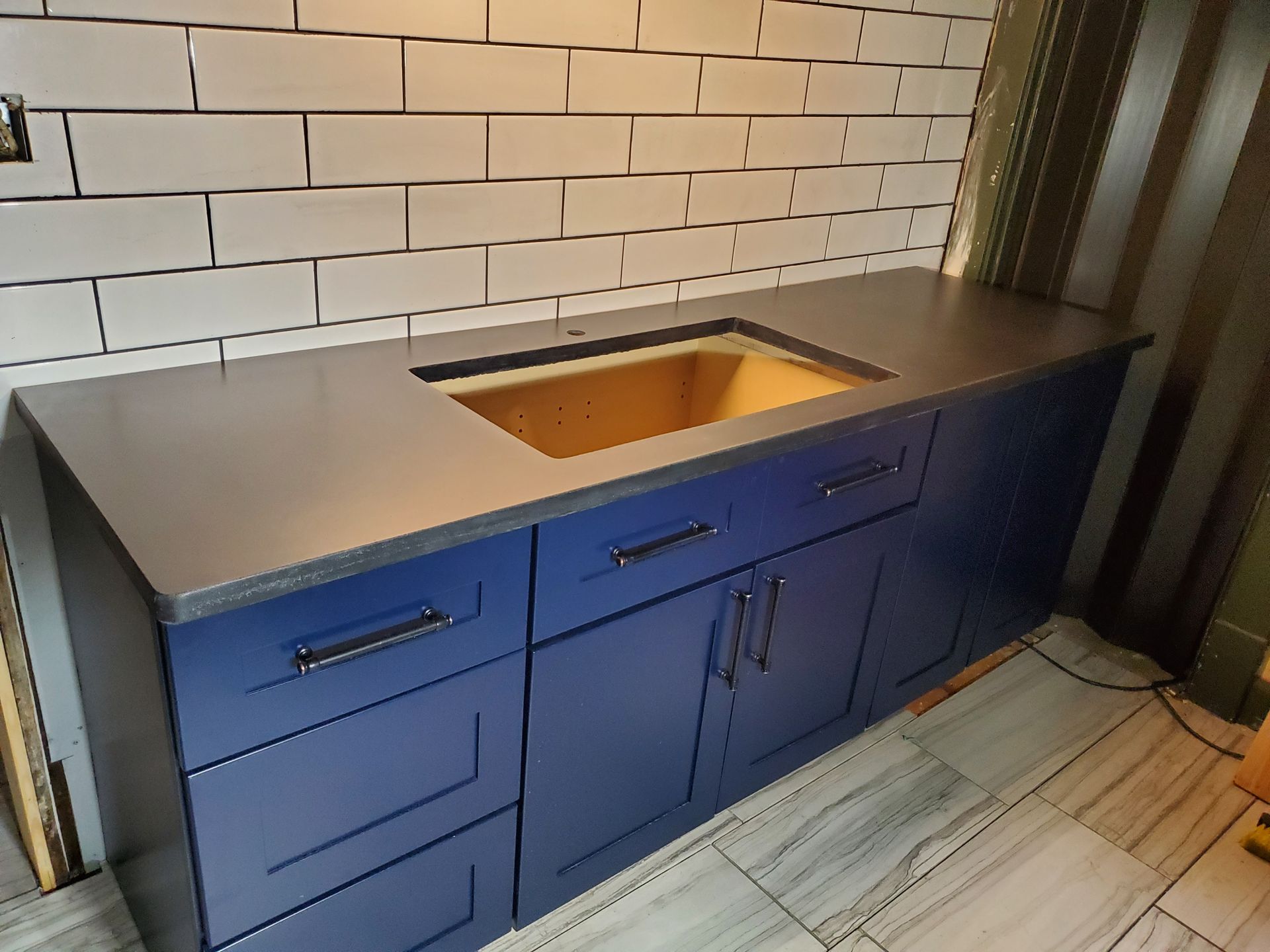 A new dark countertop with a cutout for a sink sits atop dark blue kitchen cabinets against a white subway tile backsplash.