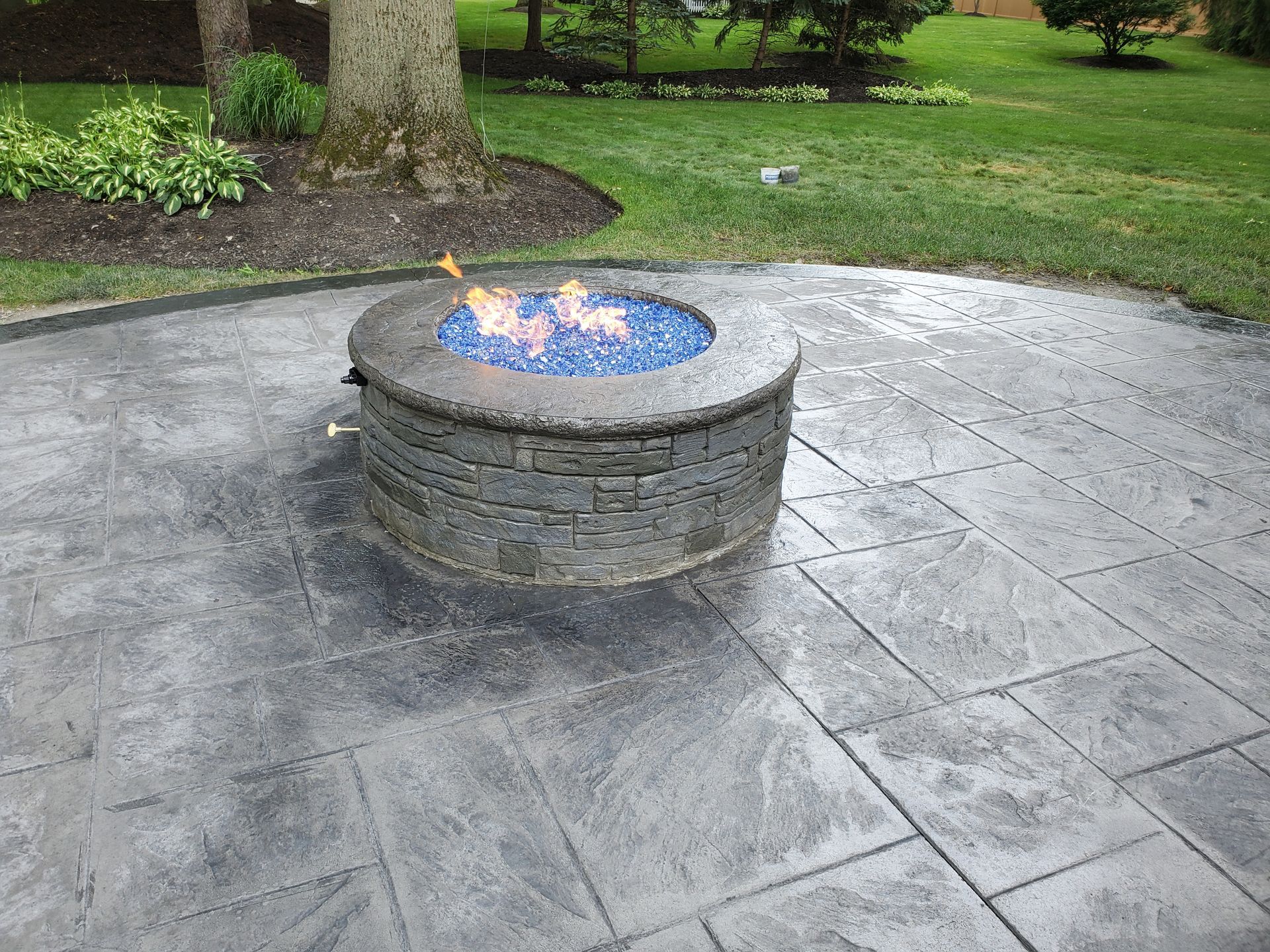 A circular stone fire pit with blue glass fire rocks and flames, centered on a gray stamped concrete patio in a yard.