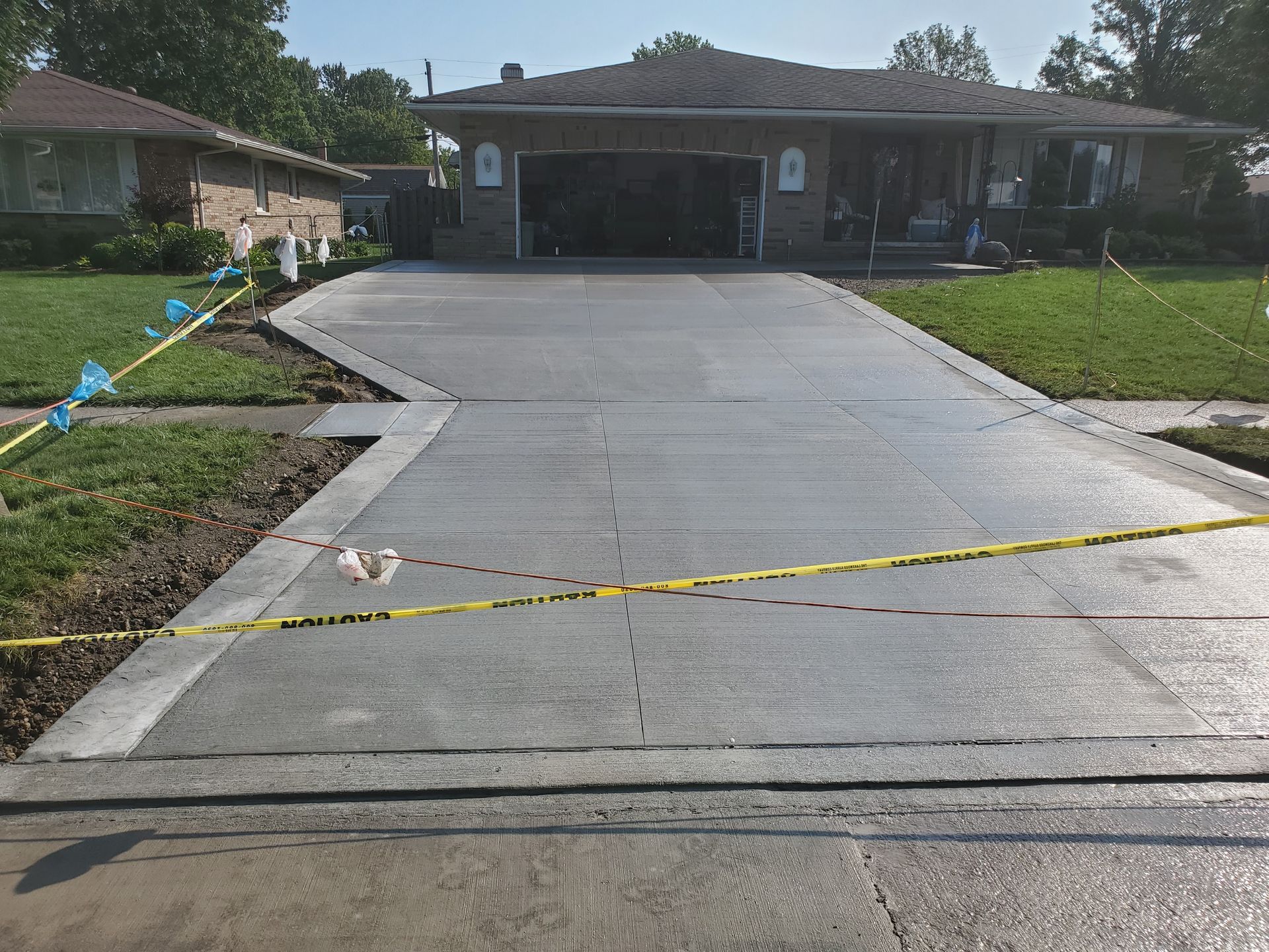 A freshly poured, smooth grey concrete driveway leading to a house garage, cordoned off by yellow caution tape.