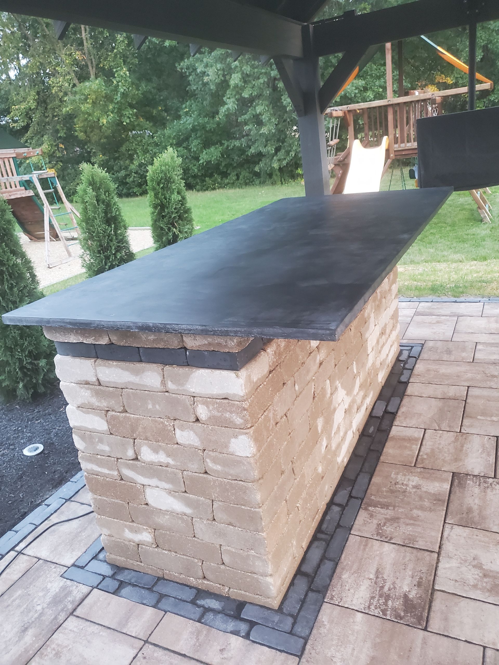 A brick outdoor bar with a dark, smooth countertop sits on a stone patio under a wooden structure in a backyard.