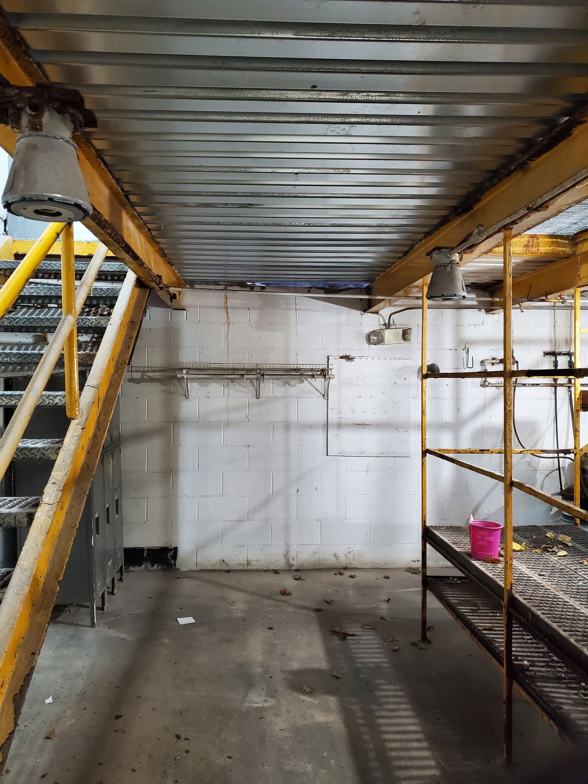 A view under a metal staircase in a room with a concrete floor, white painted walls, and metal scaffolding.