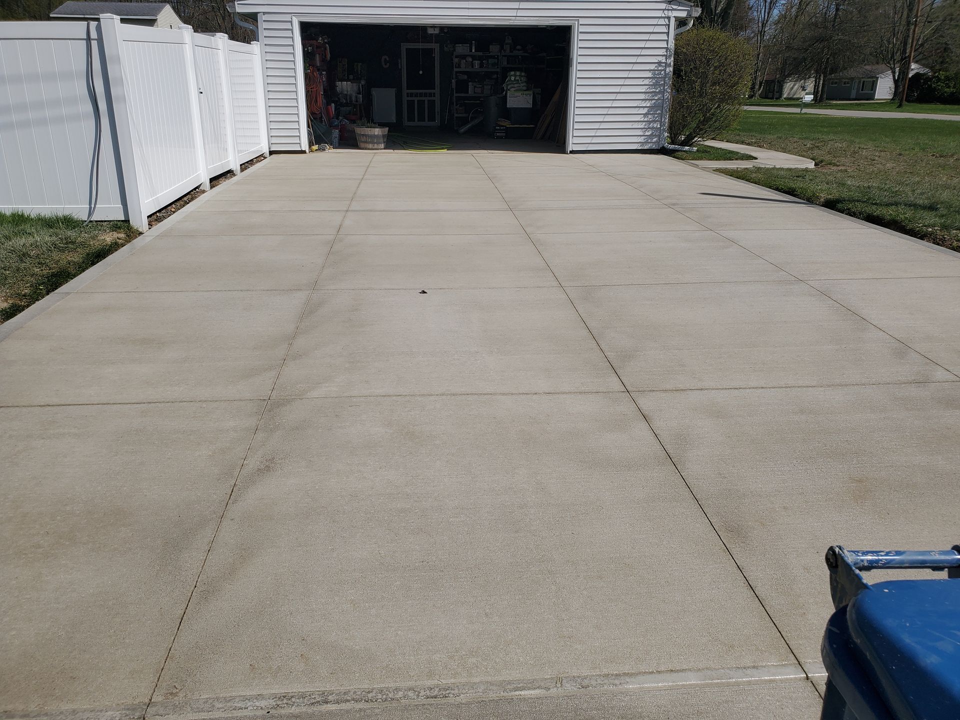 A concrete driveway leading to a white garage, flanked by a white fence on the left and a lawn on the right.