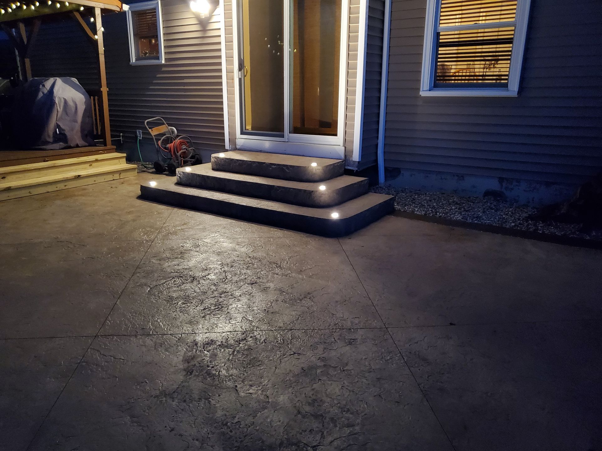 A backyard patio at night featuring stone steps leading to a sliding glass door, illuminated by integrated step lights.