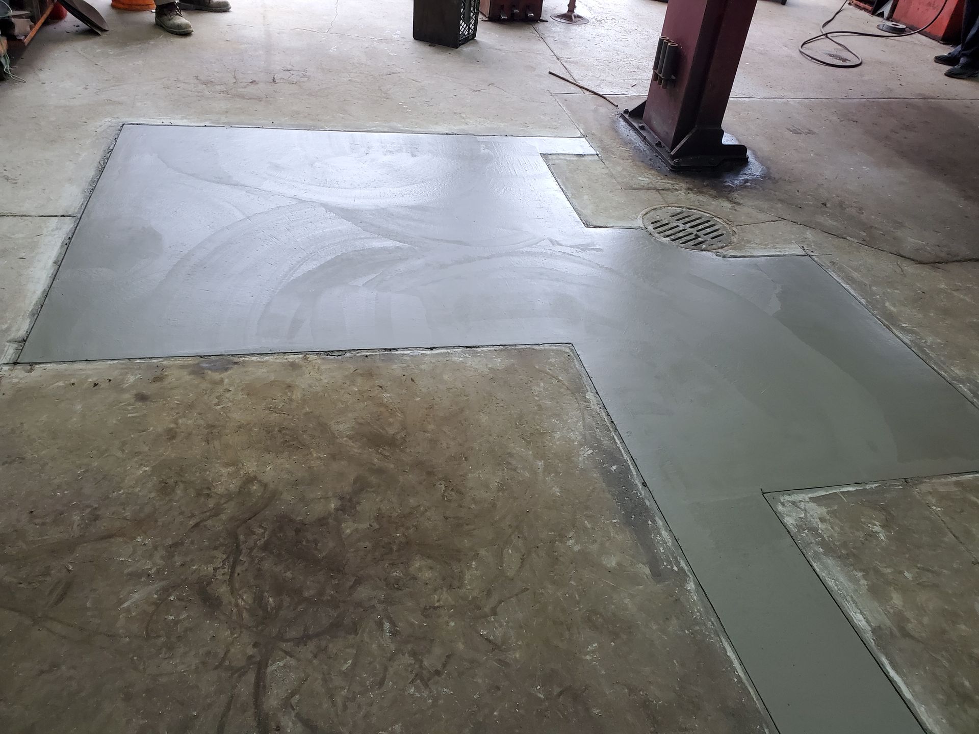 A freshly poured concrete patch on a warehouse floor, filling an L-shaped cutout in the existing rough concrete.