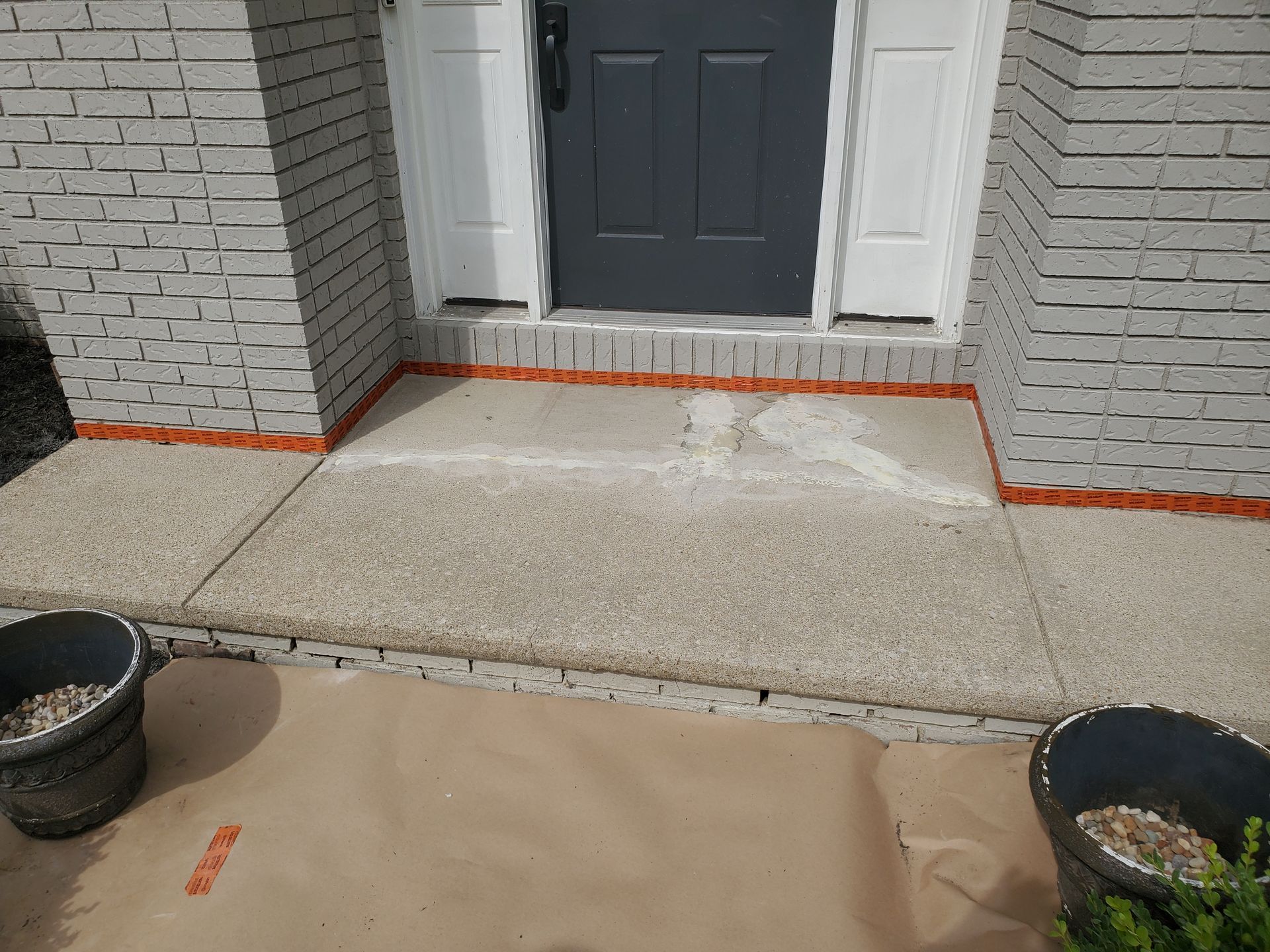 A gray-bricked home entryway featuring an orange-taped base, a gray door, and a partially cleaned concrete step.