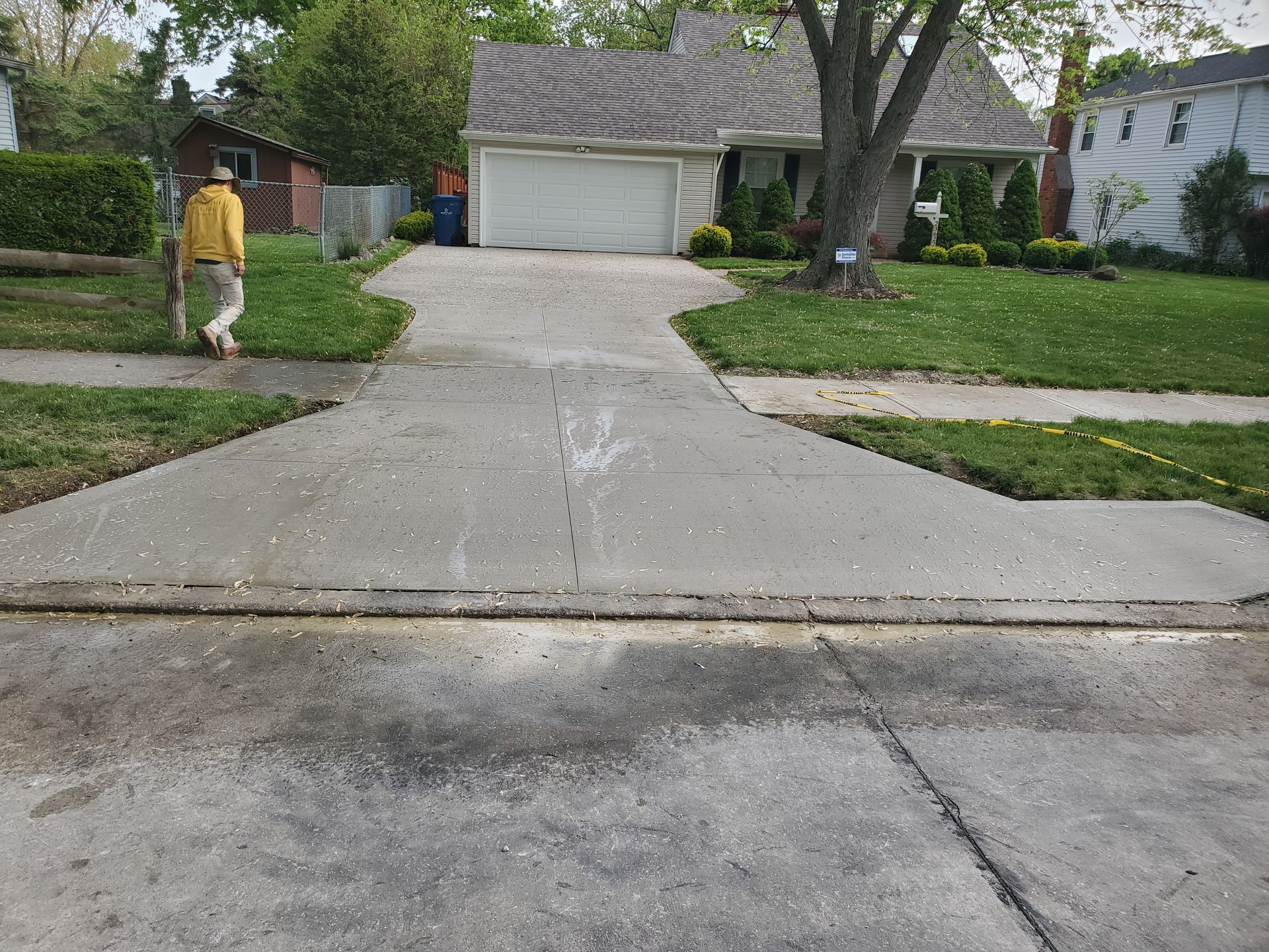A person in a yellow jacket walks up a concrete driveway toward a house with a white garage door.