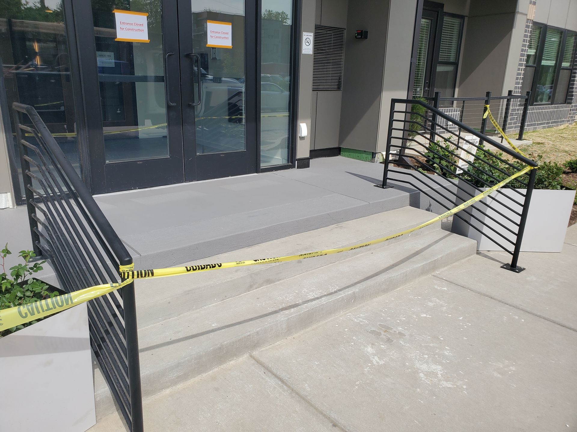 Entrance to a building with concrete stairs blocked by yellow caution tape, restricting access to the glass doors.