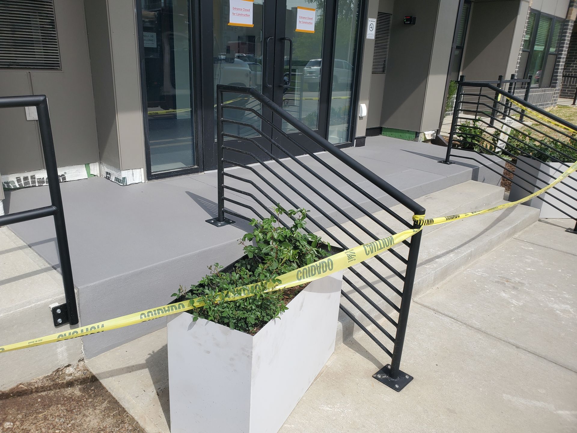 A building entrance with a concrete porch and stairs, featuring black metal railings blocked by yellow caution tape.