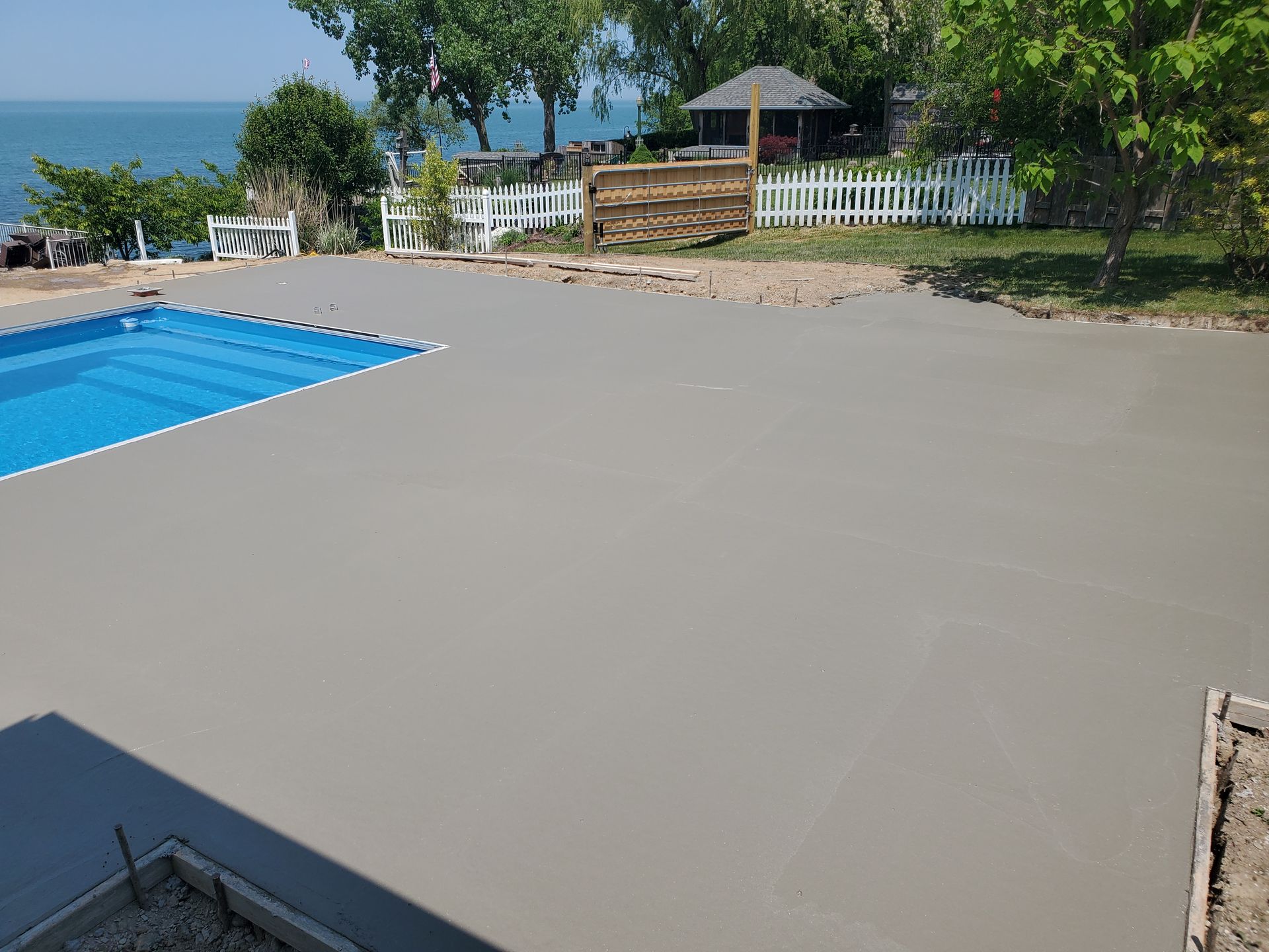 A newly poured concrete pool deck next to a blue swimming pool, overlooking a lake with trees and a white fence.