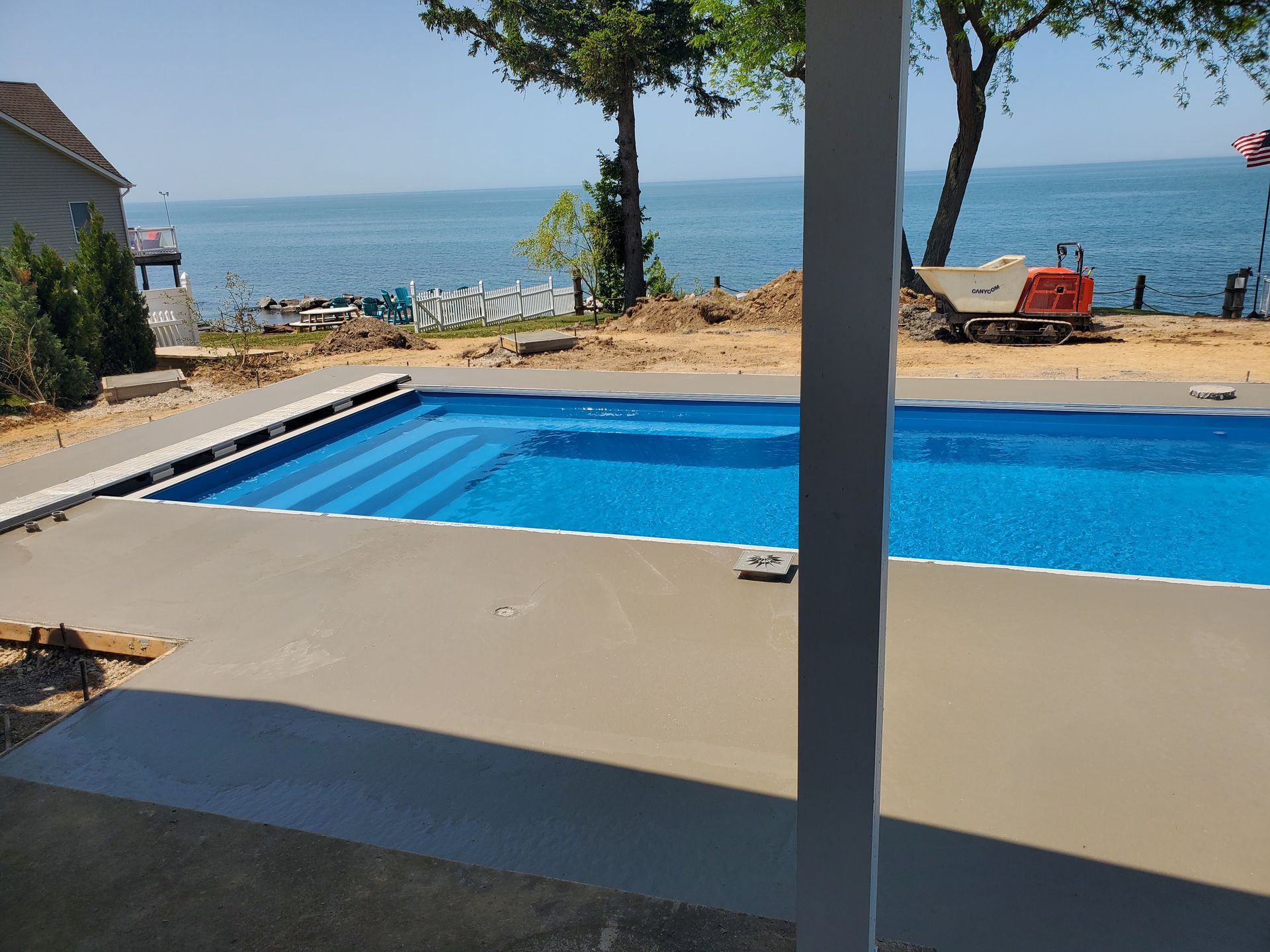 A rectangular blue swimming pool surrounded by a new concrete deck, with a view of a lake and trees in the background.