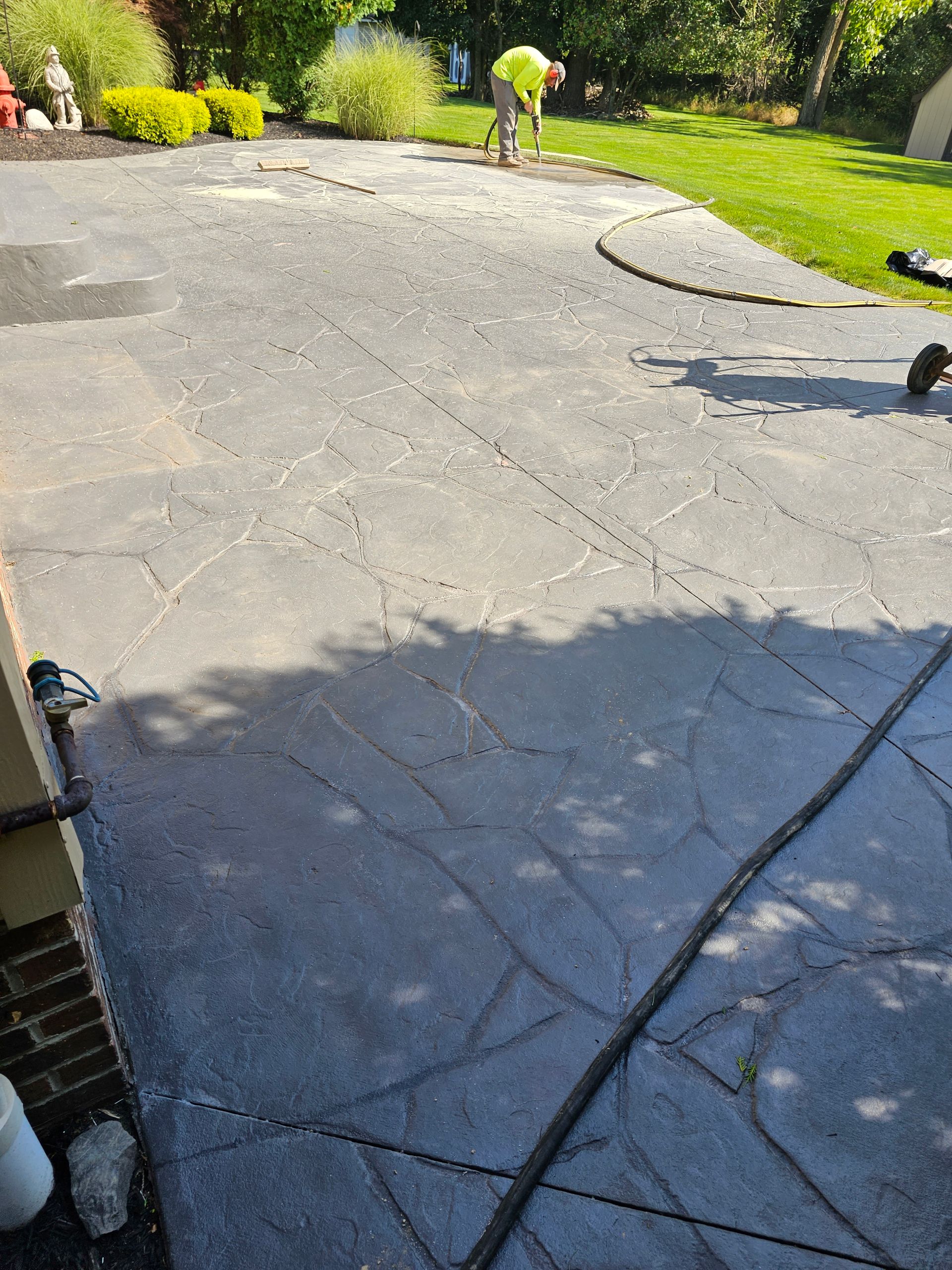 A person in a high-visibility vest works on a large, cracked asphalt patio in a residential yard.