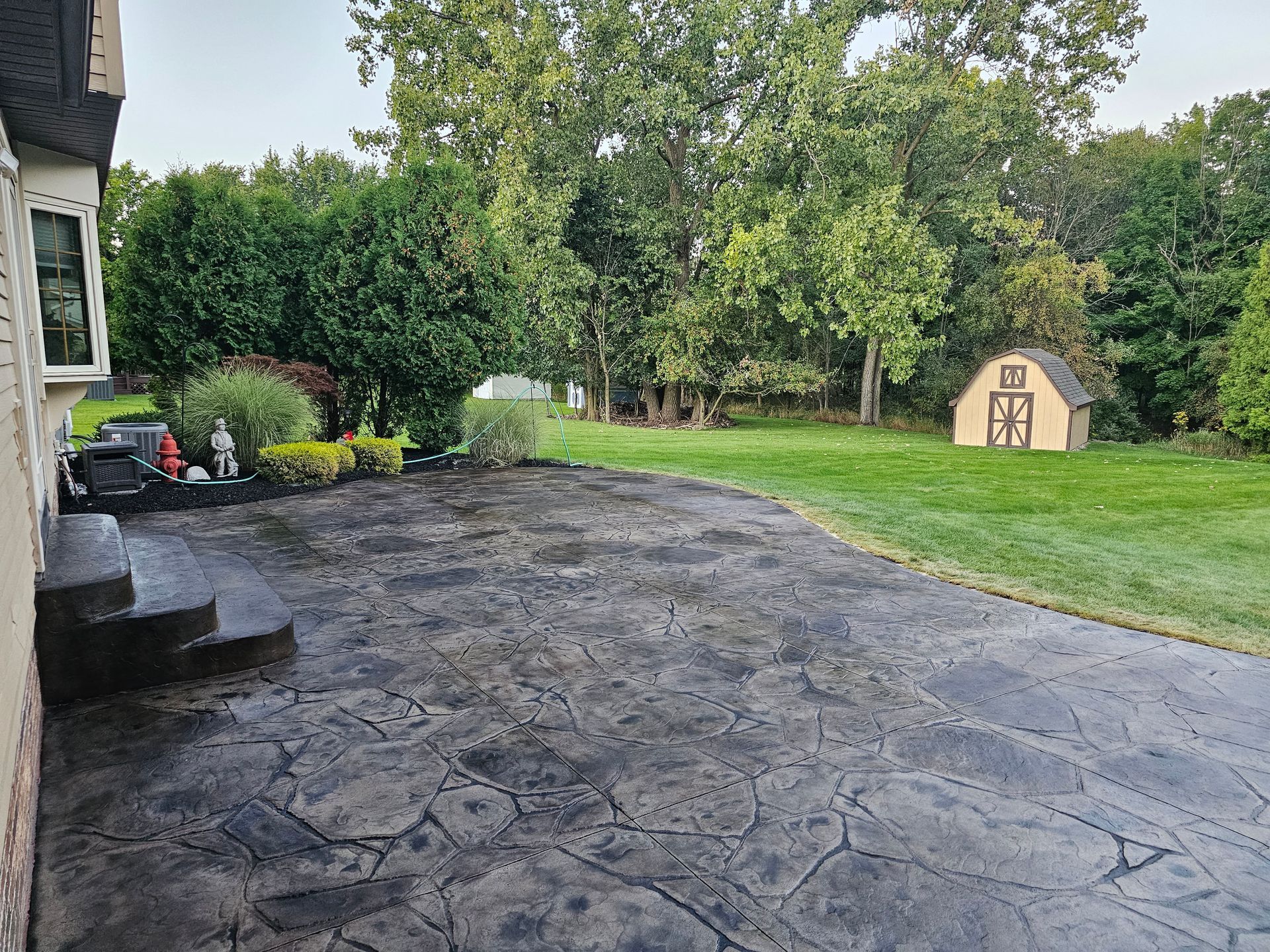 A patio with dark flagstone-style concrete leads to a grassy backyard with large trees and a small, beige shed.