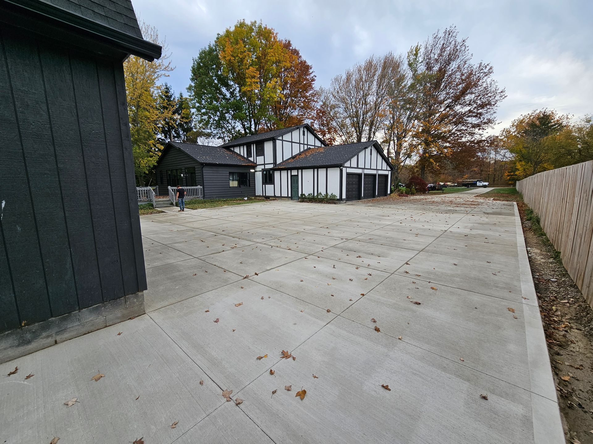 A wide concrete driveway leads to a black and white house with a multi-car garage, set against autumn trees.