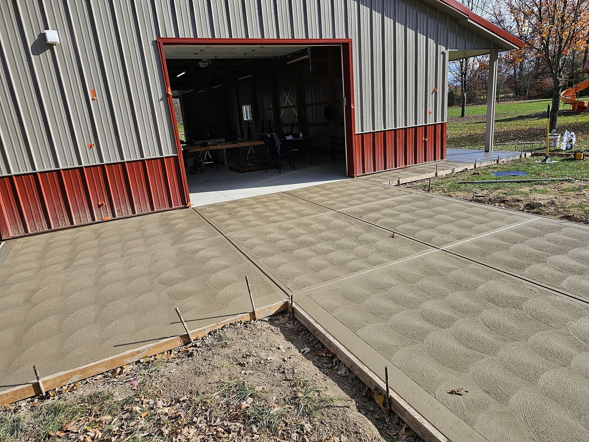 A newly poured concrete driveway apron leads into an open-bay garage building with gray siding and red trim.