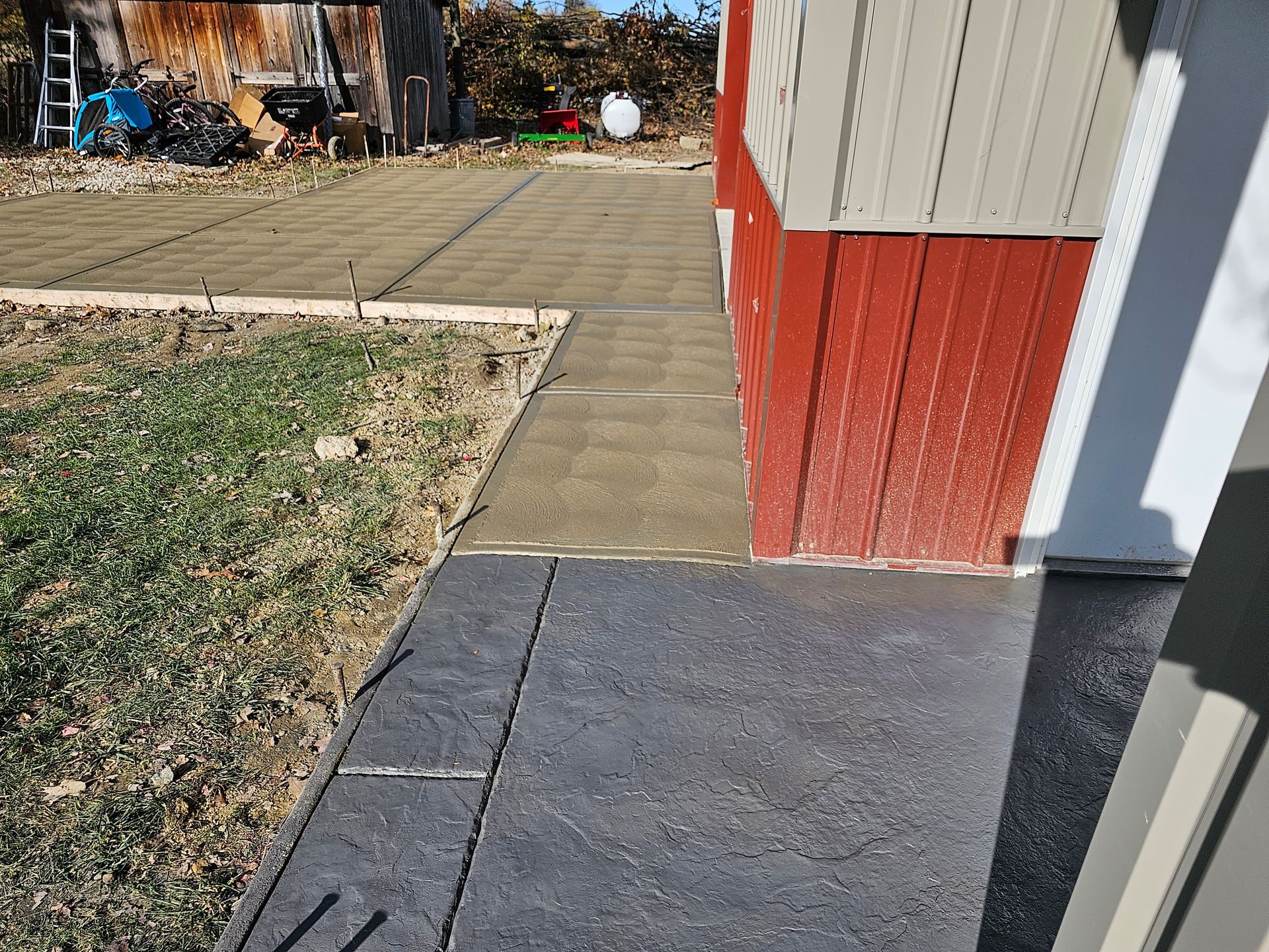 A view looking down at a transition from a dark gray paved area to lighter, square tan patio tiles beside a red building.