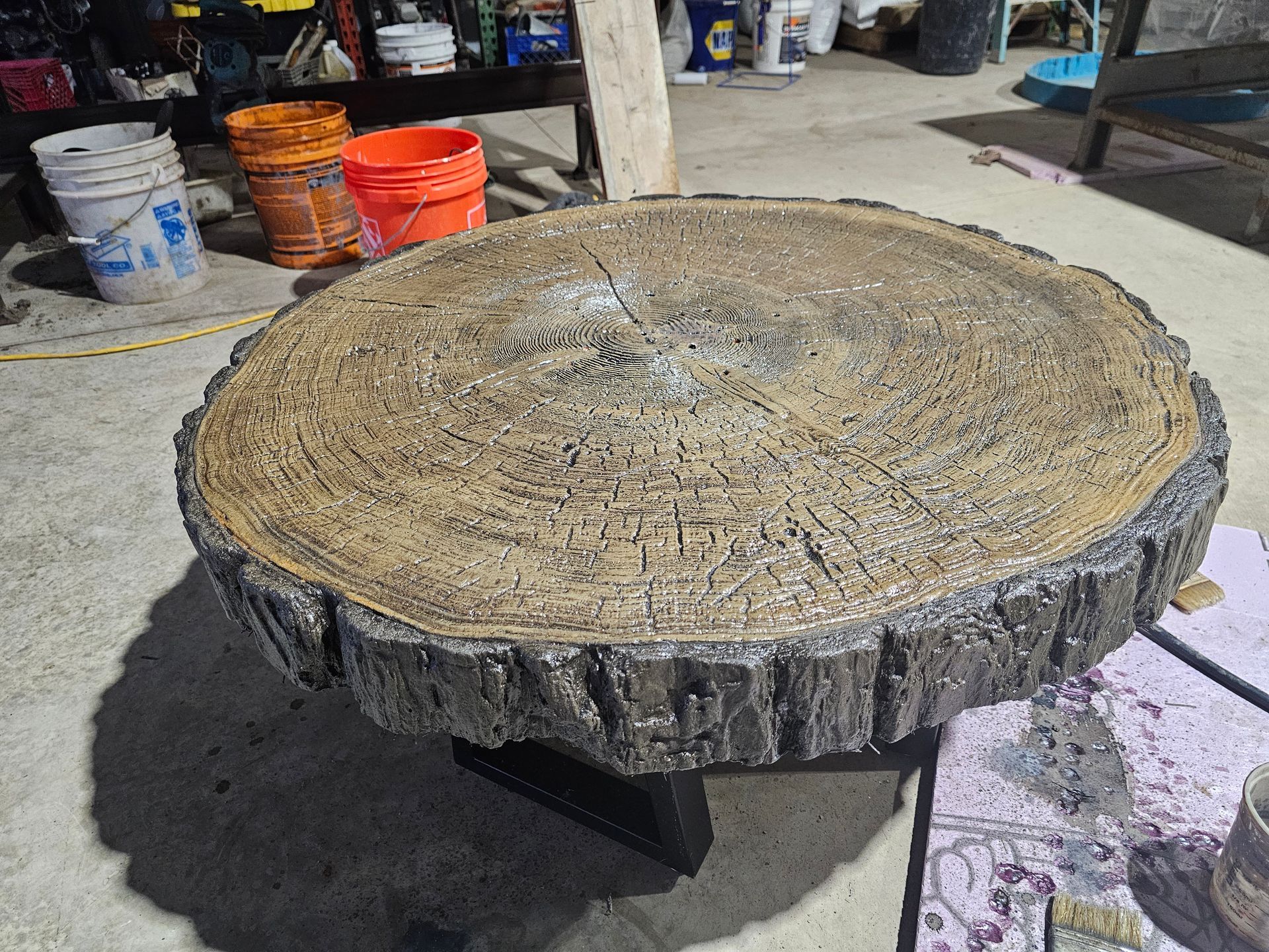 A circular table with a textured, wood-like surface and bark edges, sitting on a black base in a workshop setting.