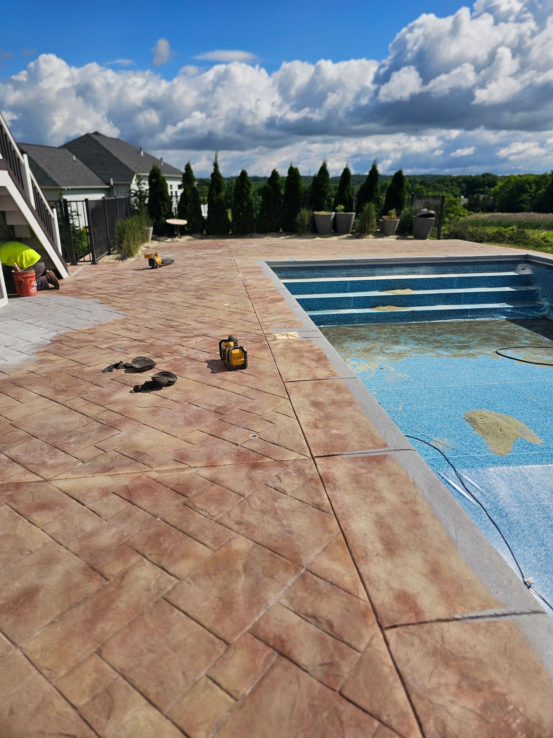 A textured, stone-patterned concrete patio surrounds a swimming pool under a bright, partly cloudy blue sky.