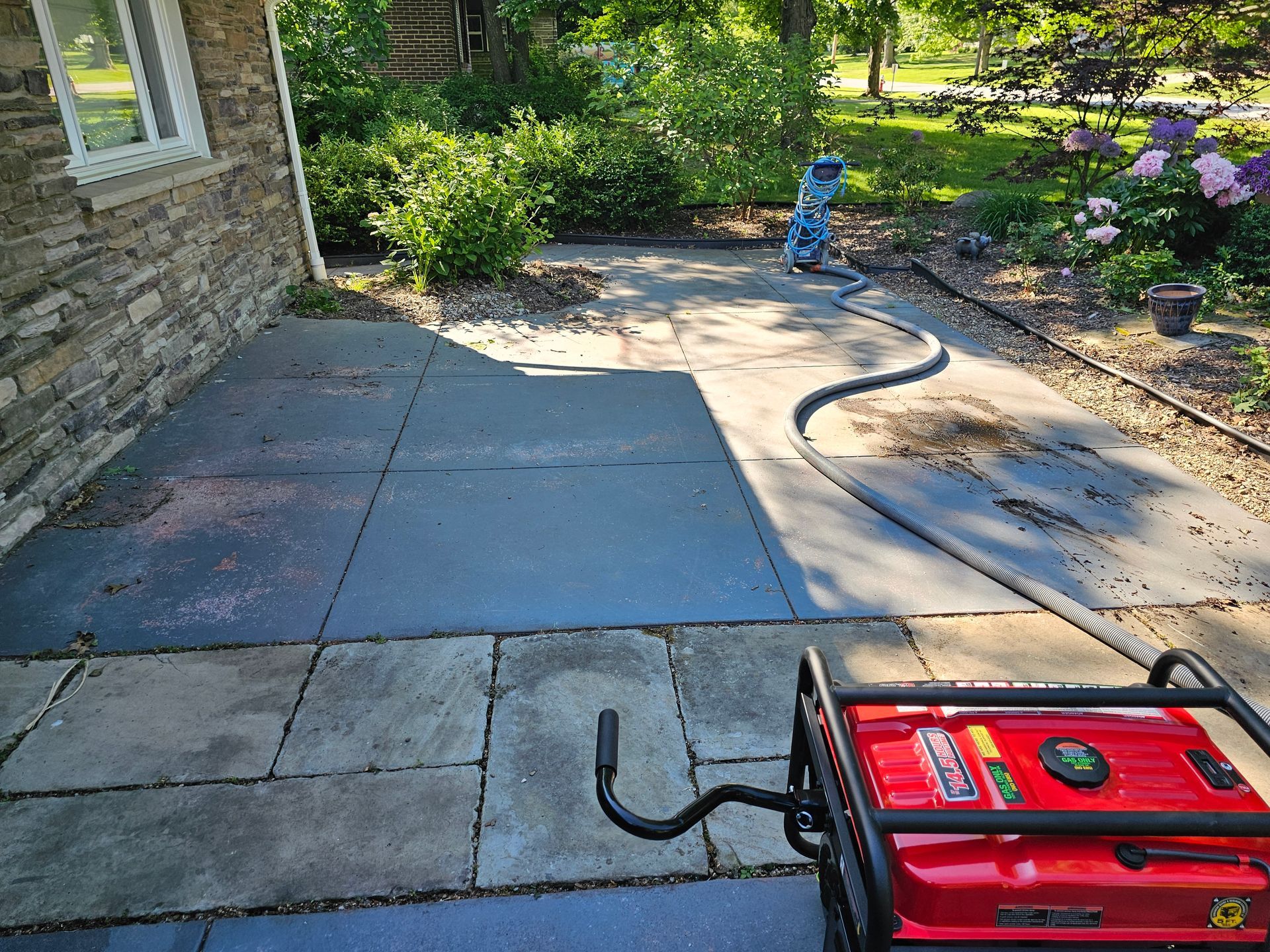 A red generator sits on a stone patio near a house, connected by a long black hose to a person cleaning the pavement.
