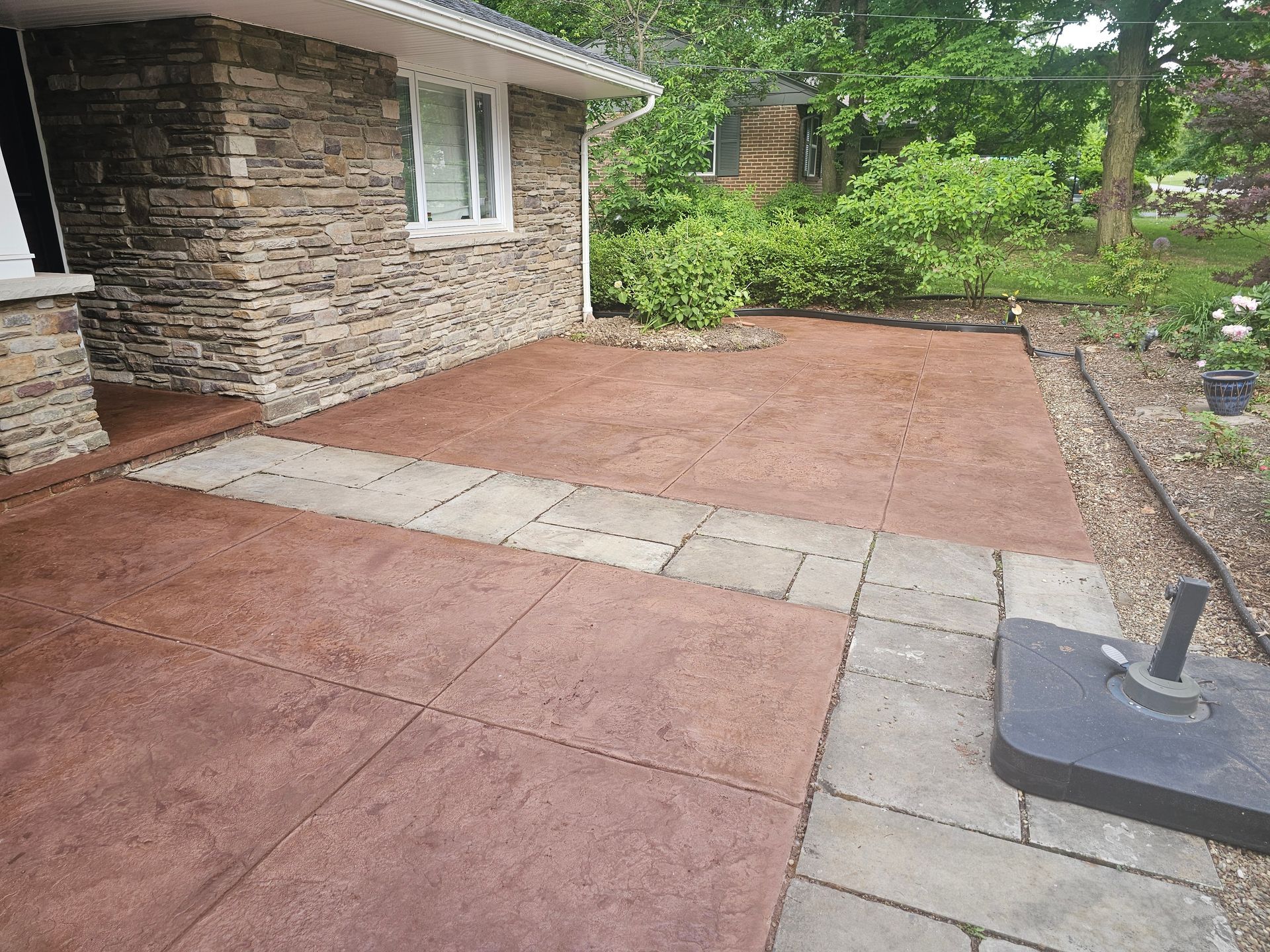 A patio with stamped reddish-brown concrete, a stone house corner, and a gray stone paver walkway leading to a garden.