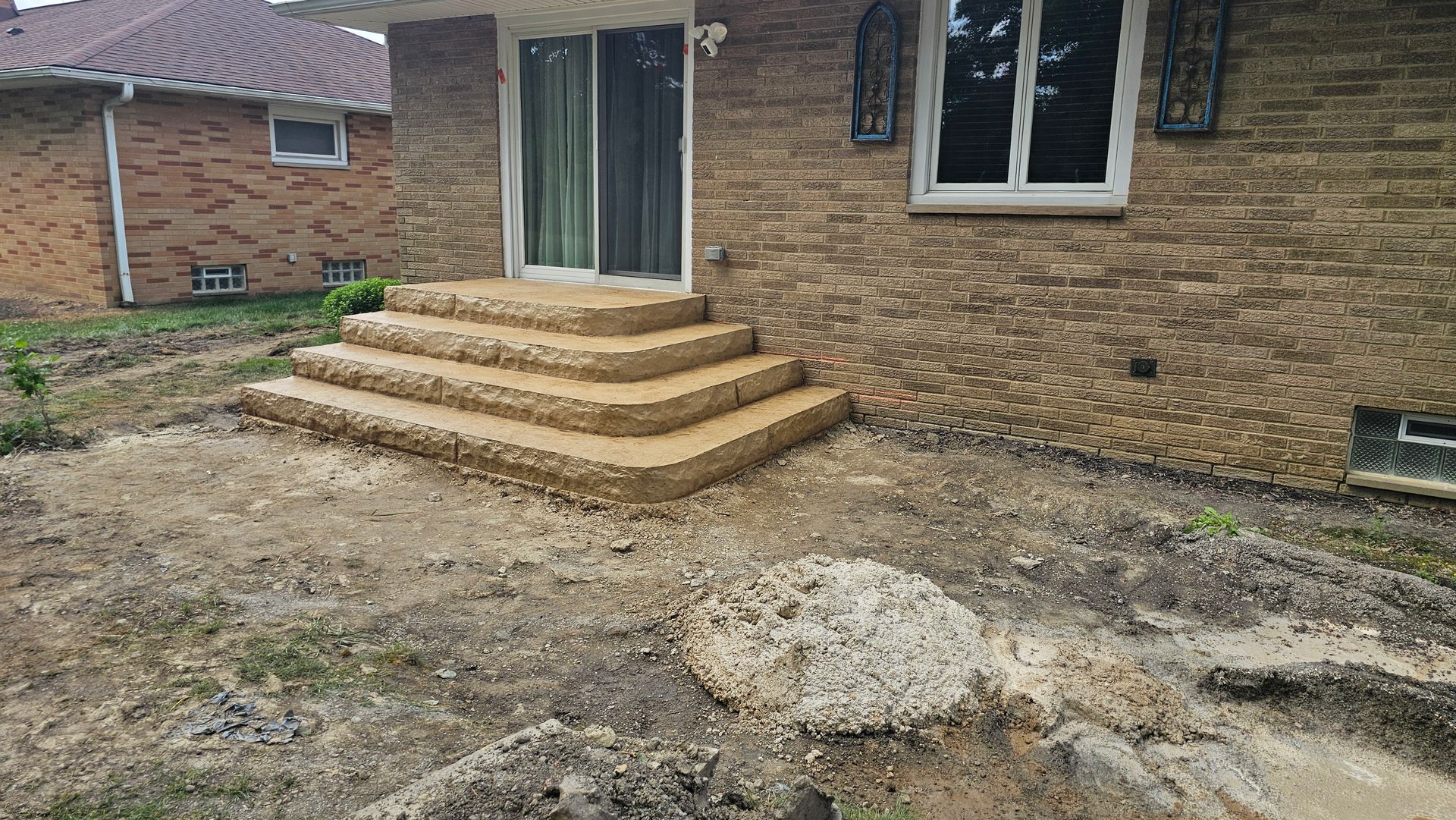 Four newly built, tan-colored concrete steps leading up to a sliding glass door on the side of a brick house.