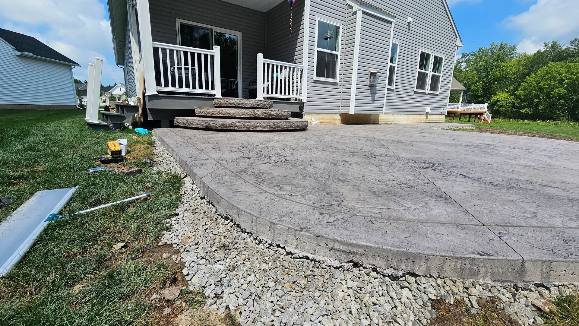 A grey stamped concrete patio with stone steps leading up to a house deck, surrounded by a gravel border and lawn.