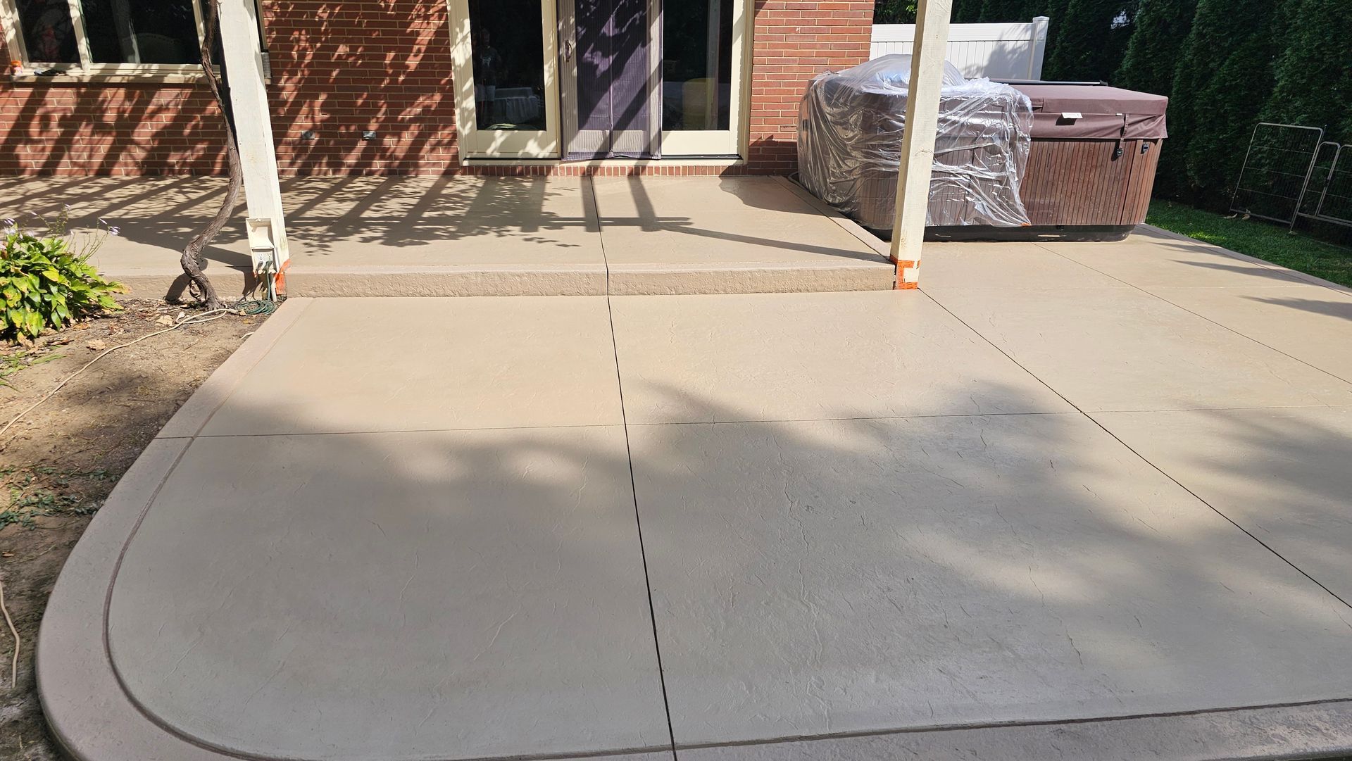A newly resurfaced beige concrete patio with a curved corner and a covered hot tub nearby, attached to a brick house.