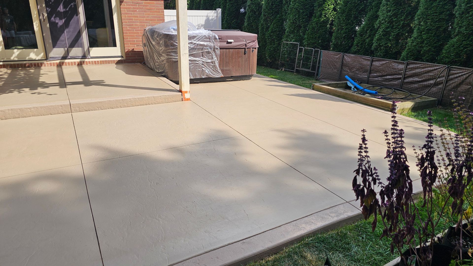 A light-colored concrete patio with expansion joints, a covered hot tub, and a landscaped backyard with a privacy fence.