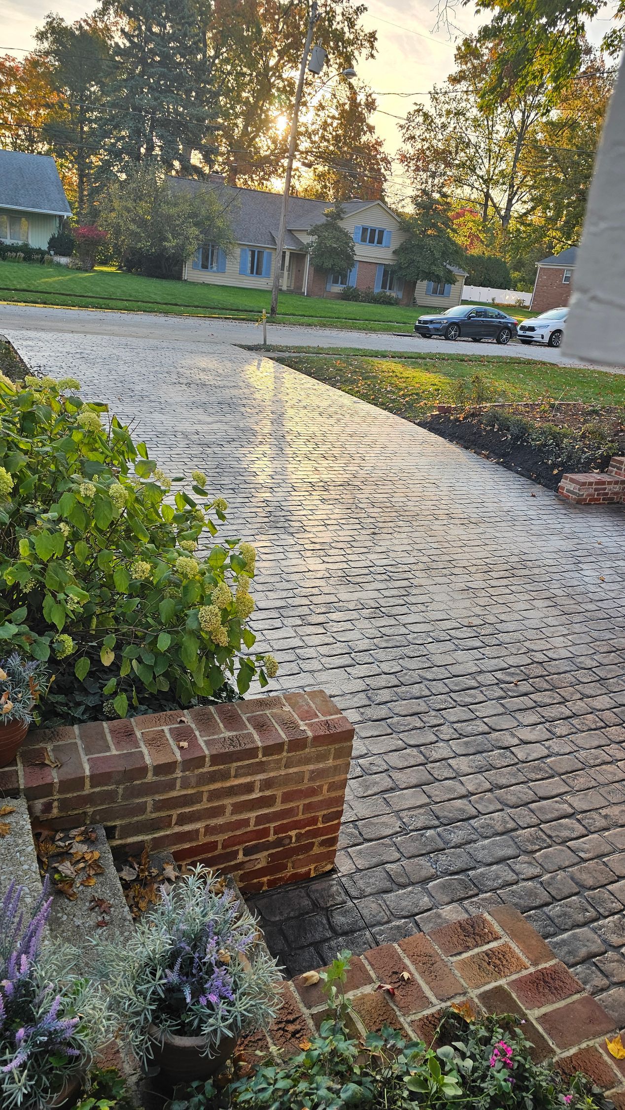 A sunlit cobblestone driveway leads toward a house in the distance, framed by brick retaining walls and garden plants.