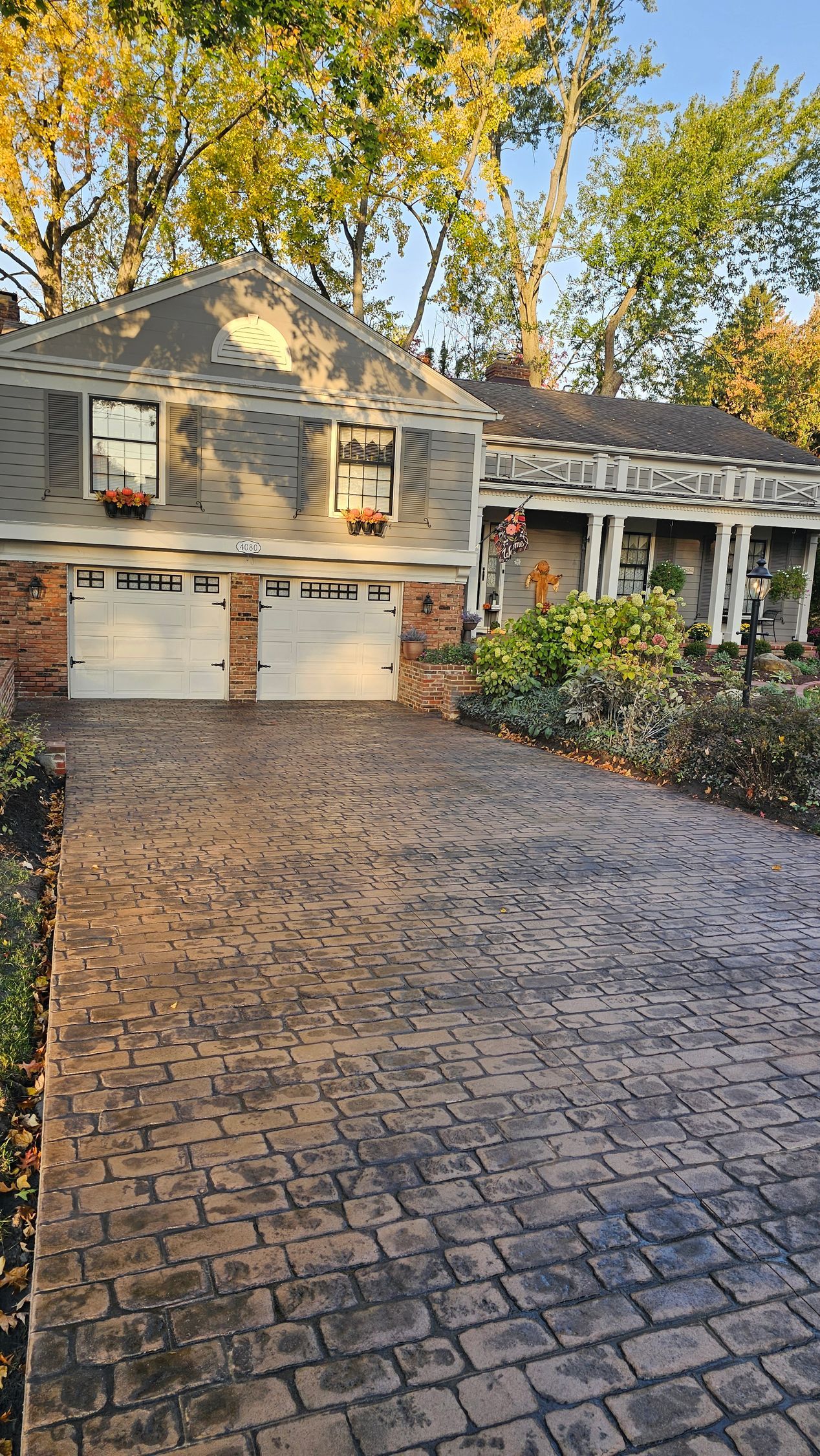 A two-story house with a textured concrete driveway and a white garage, set against autumn trees in soft sunlight.
