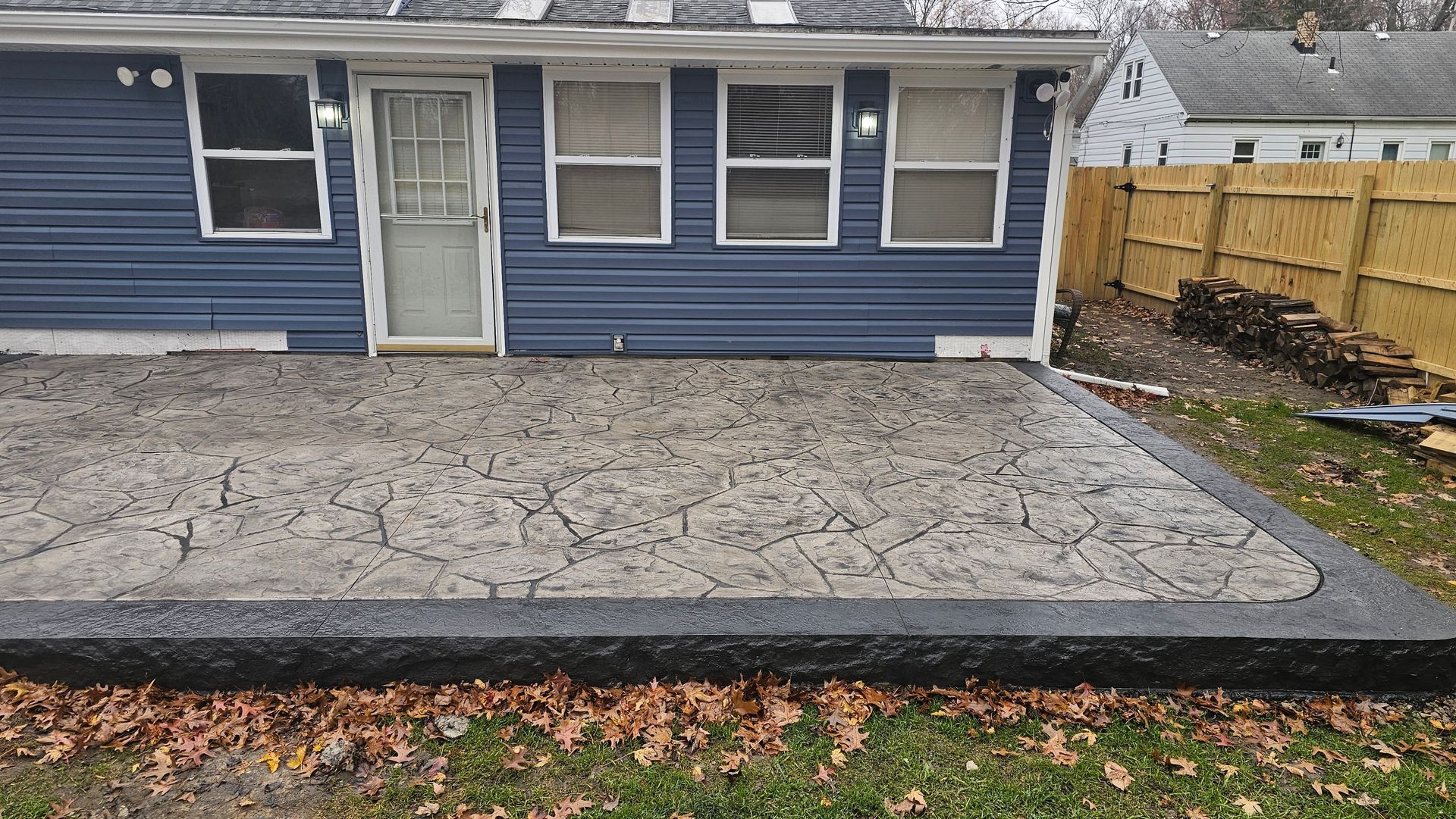 A newly poured gray stamped concrete patio with a dark border edge, situated behind a blue-sided house with a lawn.