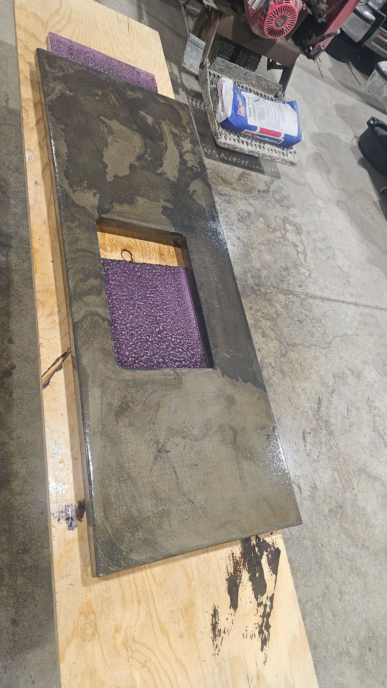 A rough concrete slab with a rectangular sink cutout sits on a plywood board in a workshop.