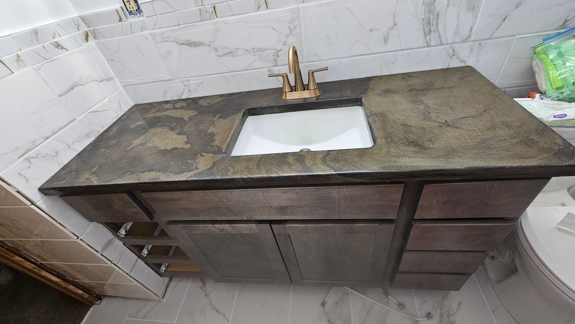A dark stone vanity countertop with a gold faucet and rectangular white sink, set against white marble-tiled walls.