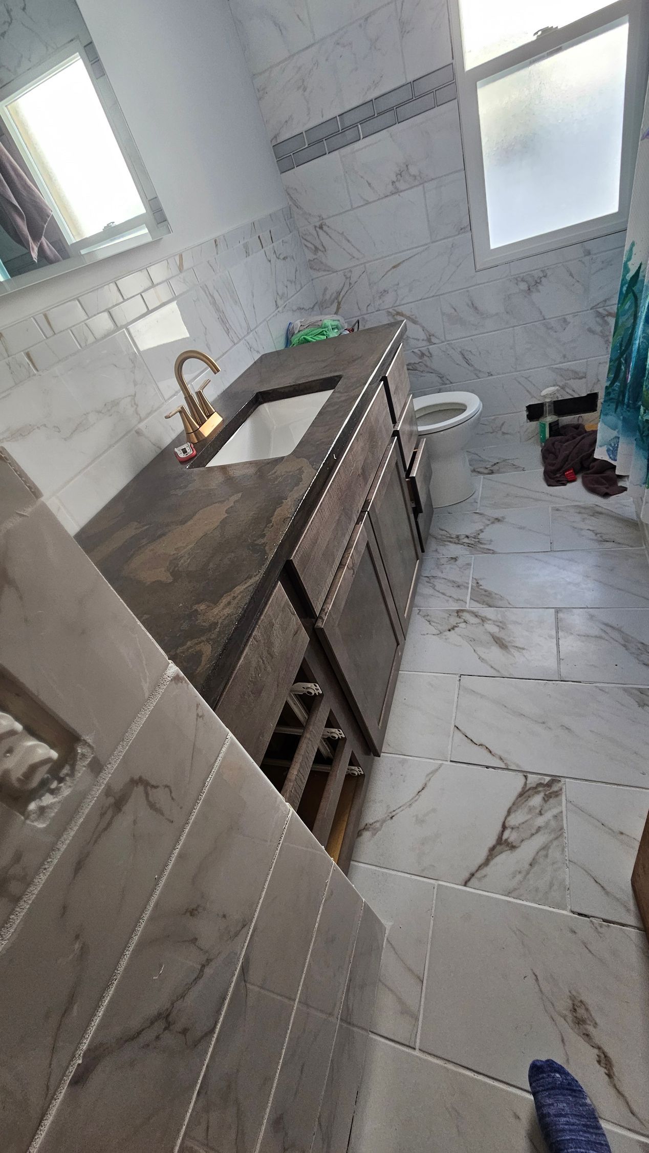 A bathroom with a dark wood vanity, white marble-style wall and floor tiles, a gold faucet, and a toilet in the corner.