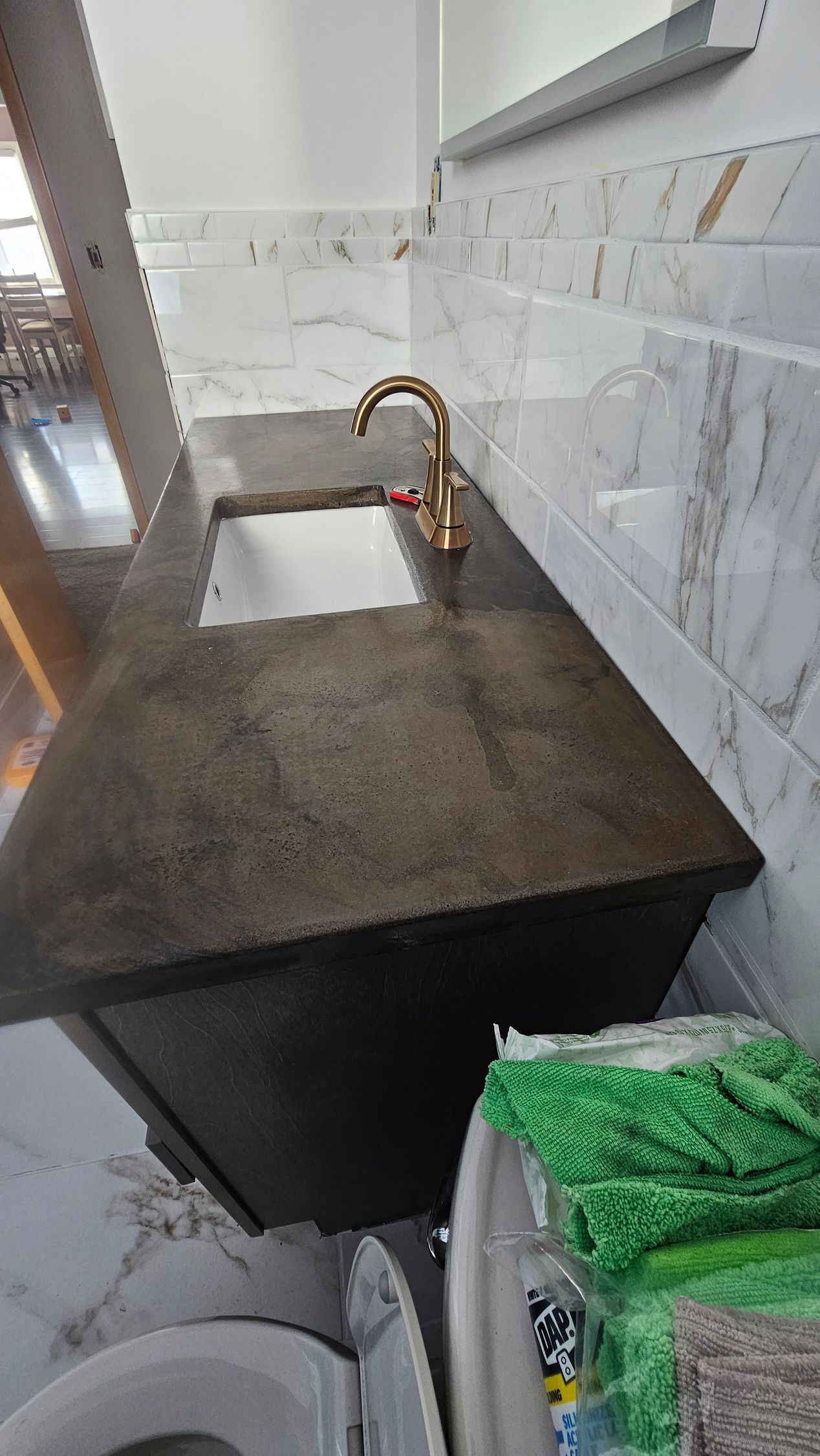 A dark concrete bathroom vanity with a white rectangular sink and a gold faucet against a white tiled wall.