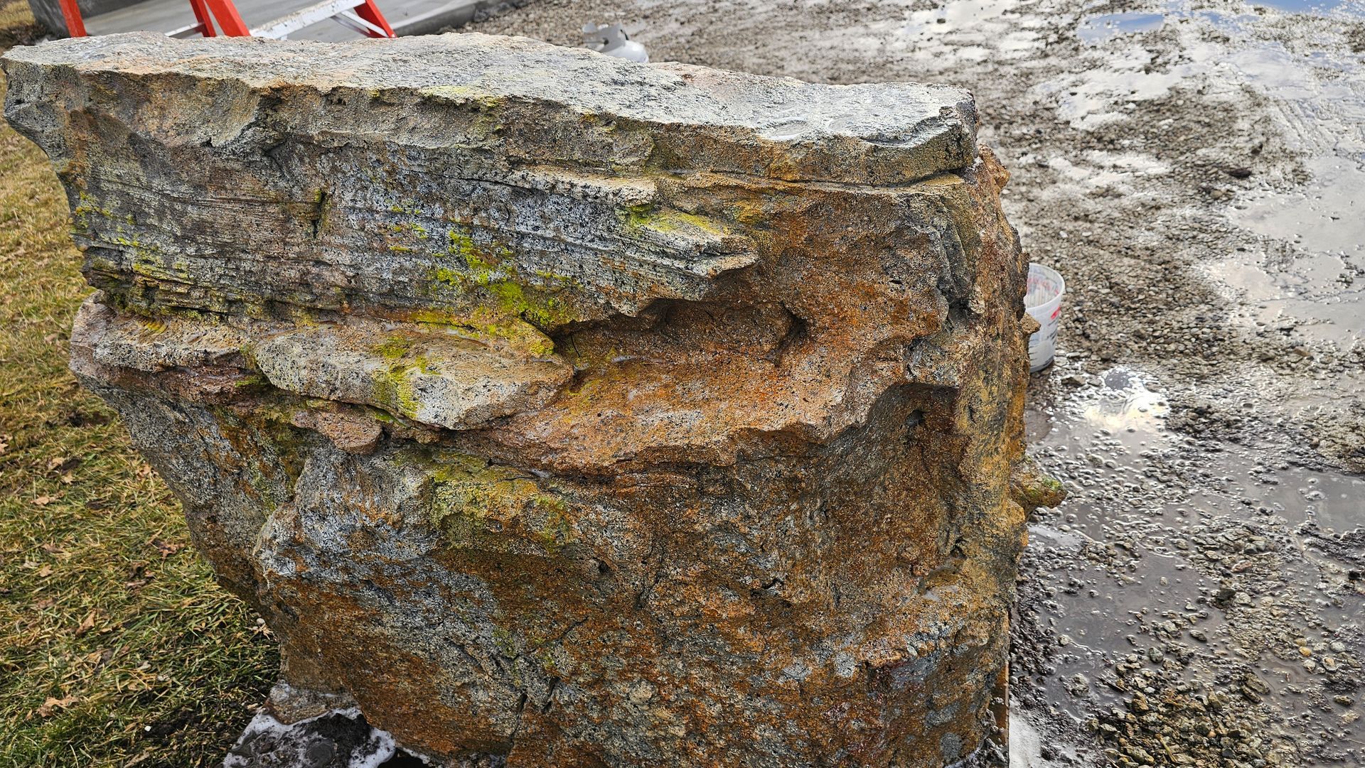 A large, weathered stone boulder with rough, textured surfaces, yellow-green moss, and rusty-brown mineral staining.