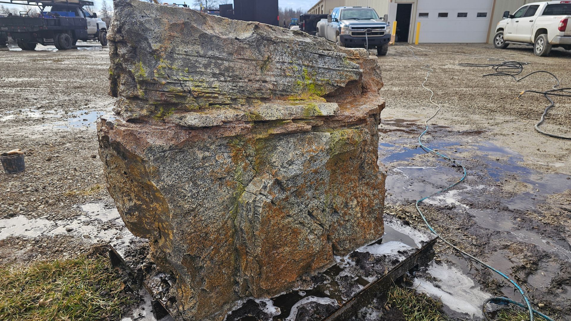 A large, rough, mossy brown stone block sits in a muddy outdoor industrial or construction lot near parked trucks.