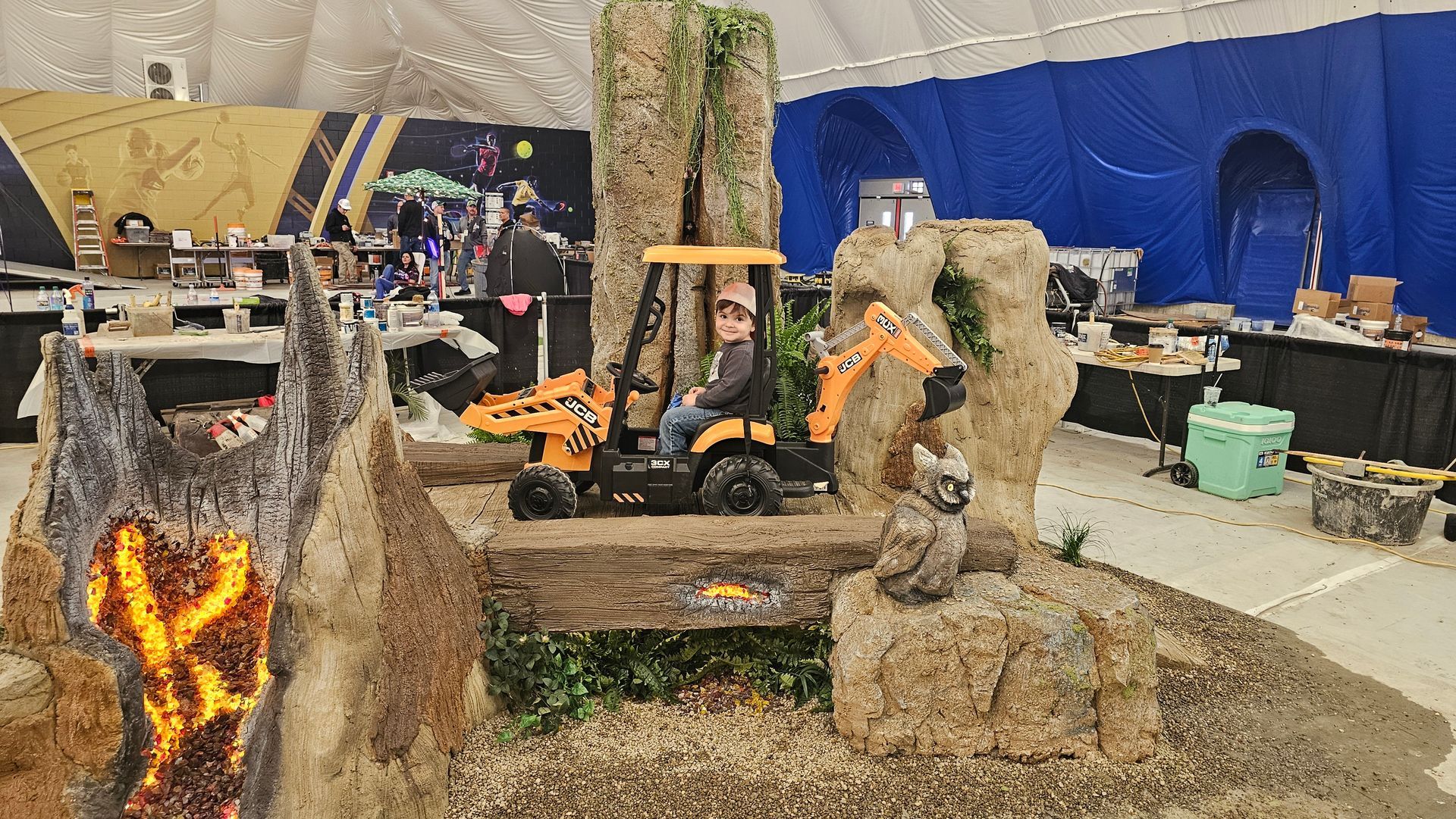 A child sits in a toy orange excavator displayed in a simulated nature exhibit with a faux volcano and rock formations.