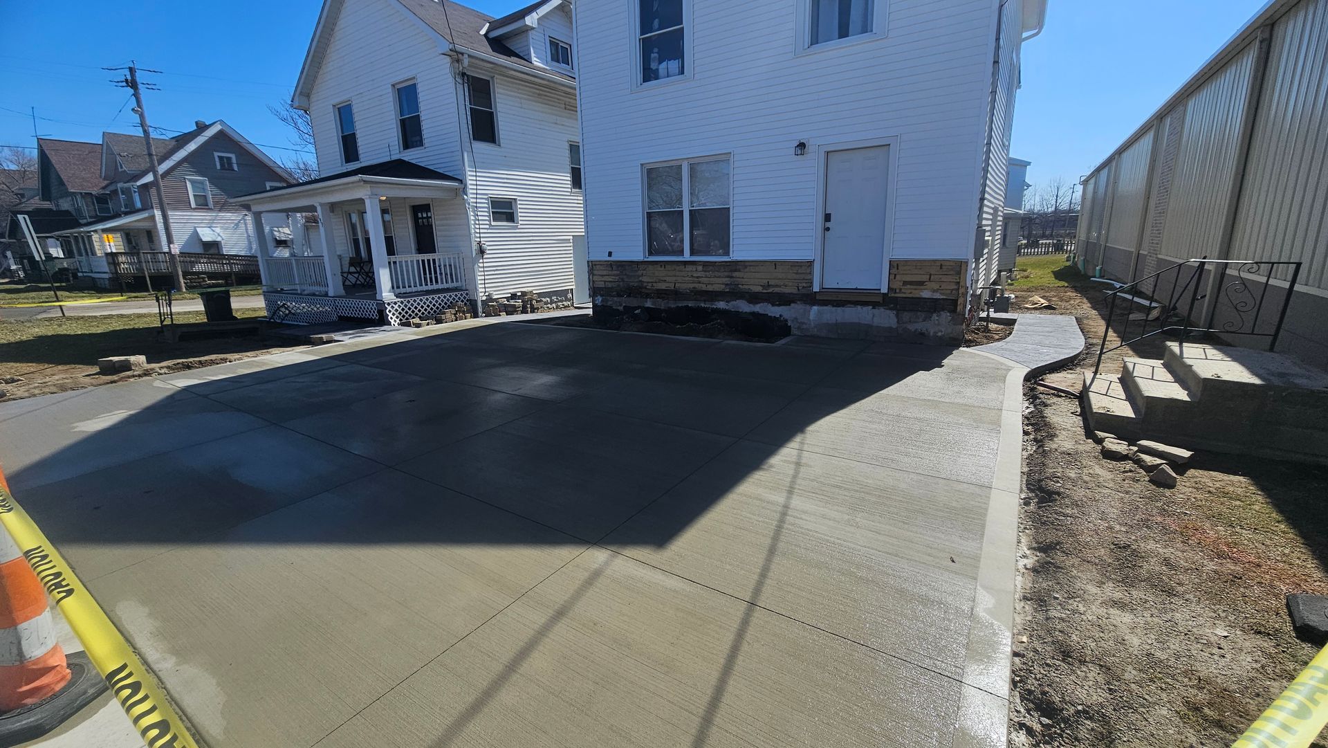 A newly poured concrete driveway between two residential houses on a sunny day, marked off by yellow caution tape.