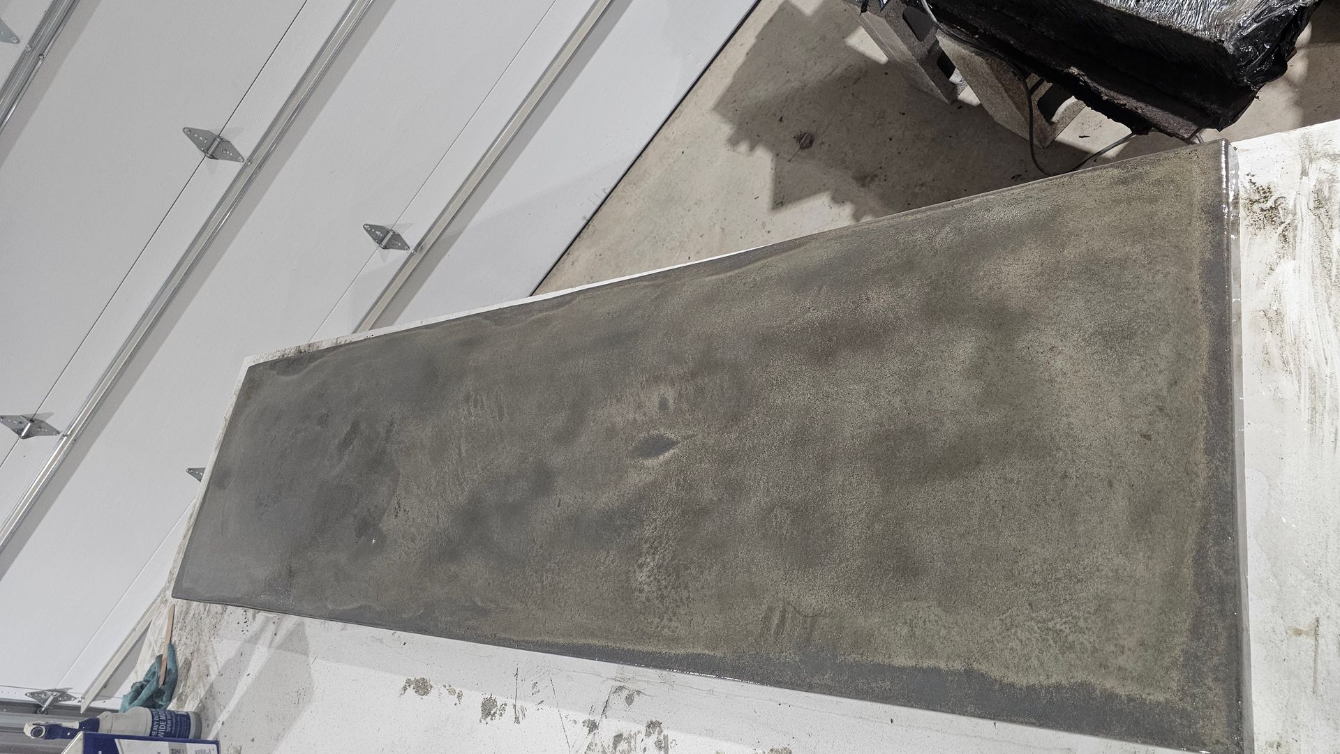 A freshly poured rectangular concrete pad sits on a white surface next to a garage door.