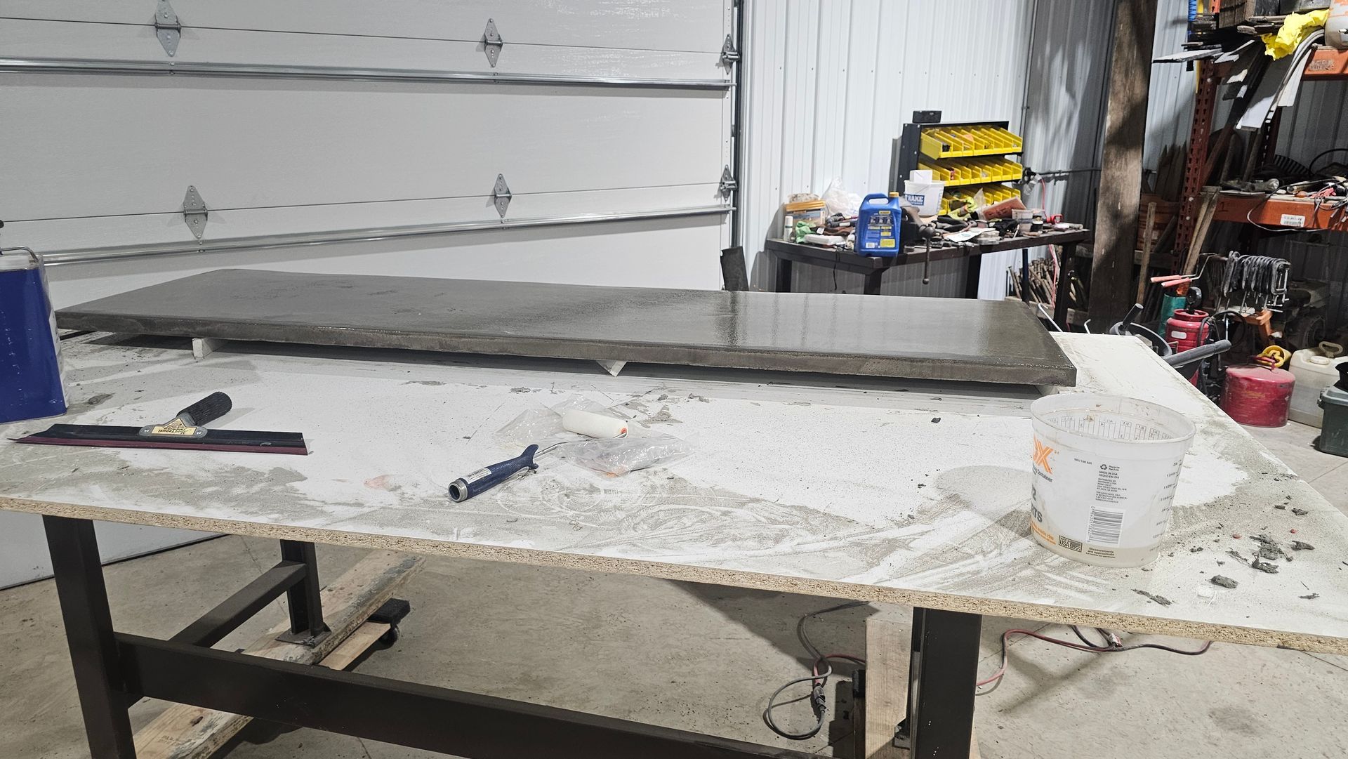 A large, dark concrete slab rests on a workbench in a cluttered garage, with a trowel and bucket nearby.