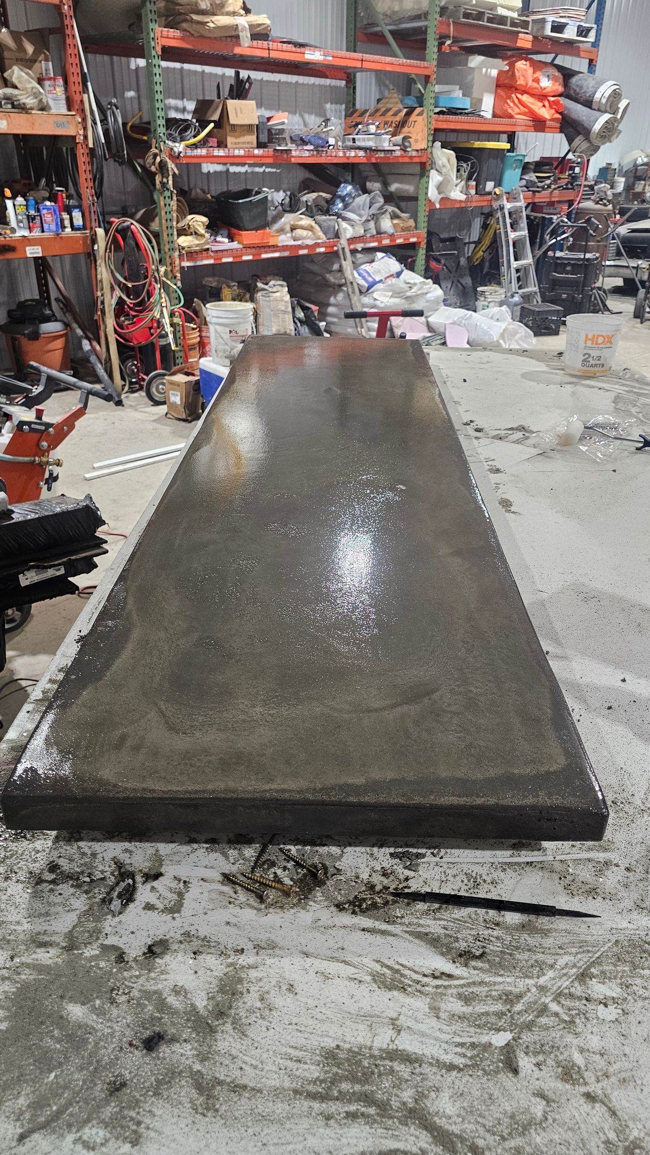 A wet, rectangular concrete slab sits on a dusty workshop floor in front of metal shelving units.