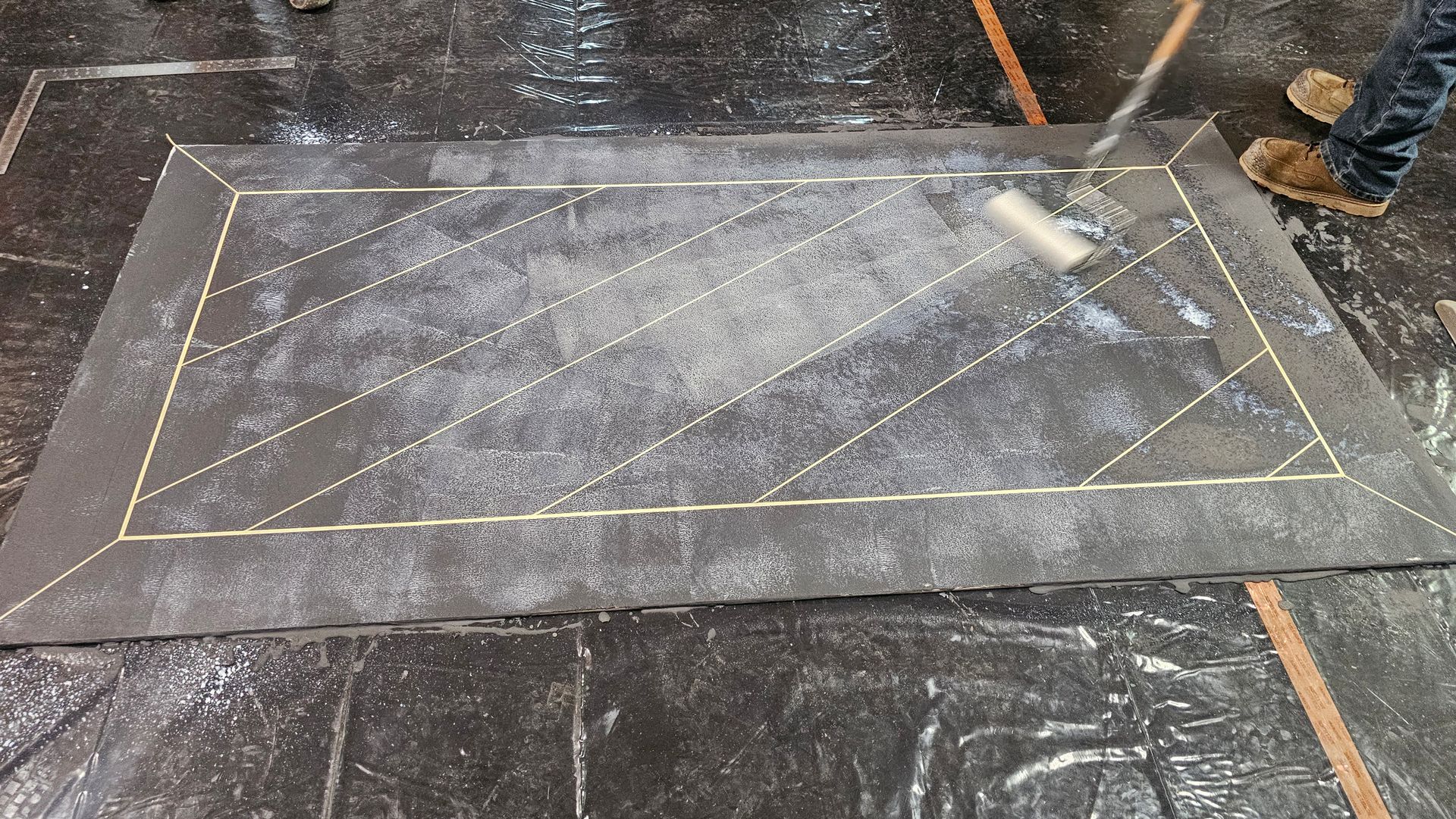 A person uses a roller to apply a light-colored liquid coating to a rectangular section of a dark floor marked with lines.