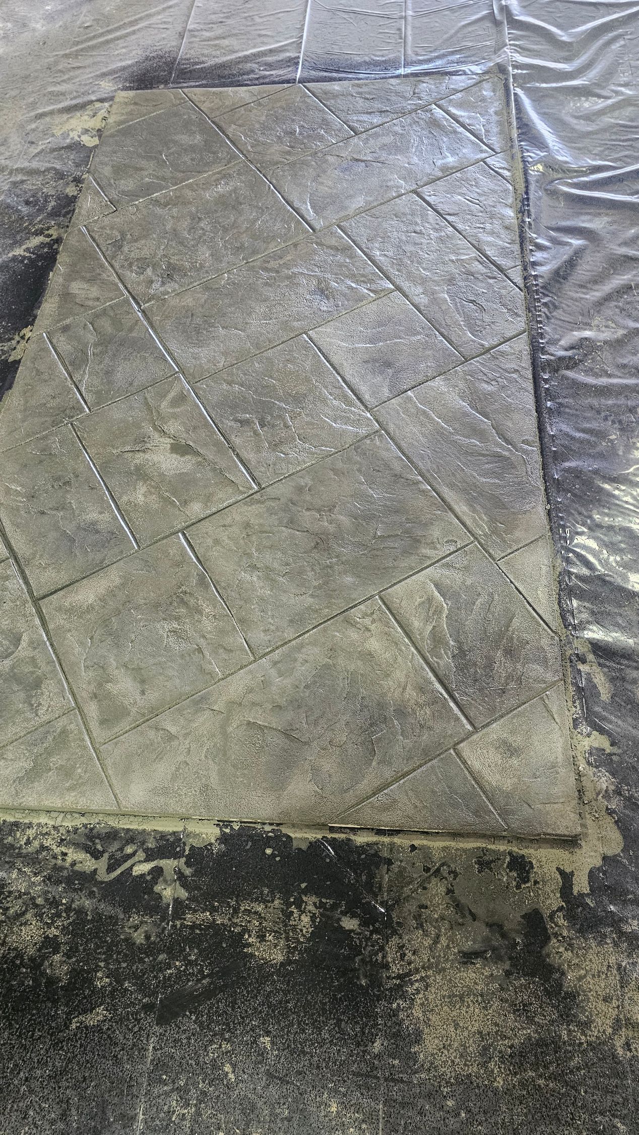 A rectangular section of stamped, grey concrete featuring a diagonal tiled pattern, set against a rough, dark ground.
